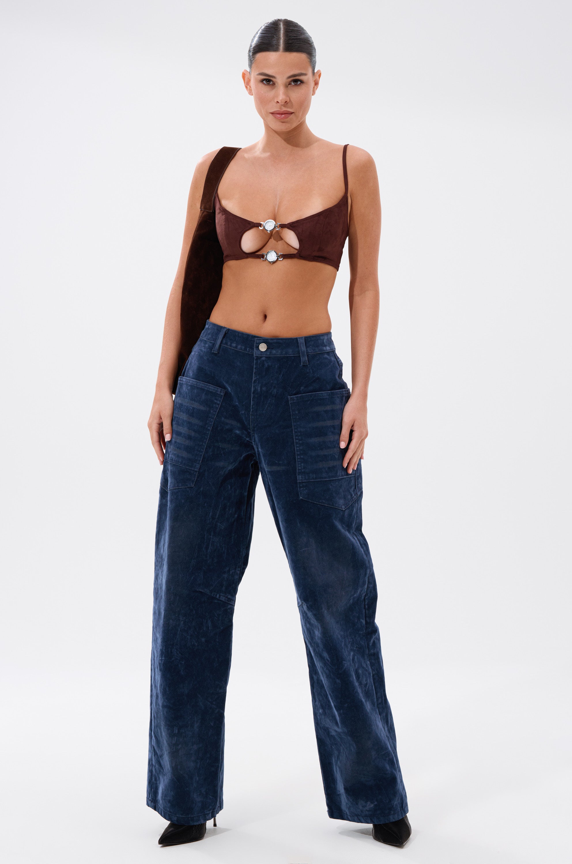 A woman poses against a white background wearing the READ BETWEEN THE LINES CLOCK BRALETTE, high-waisted wide-leg blue velvet pants, black heels, and carrying a brown shoulder bag. 7