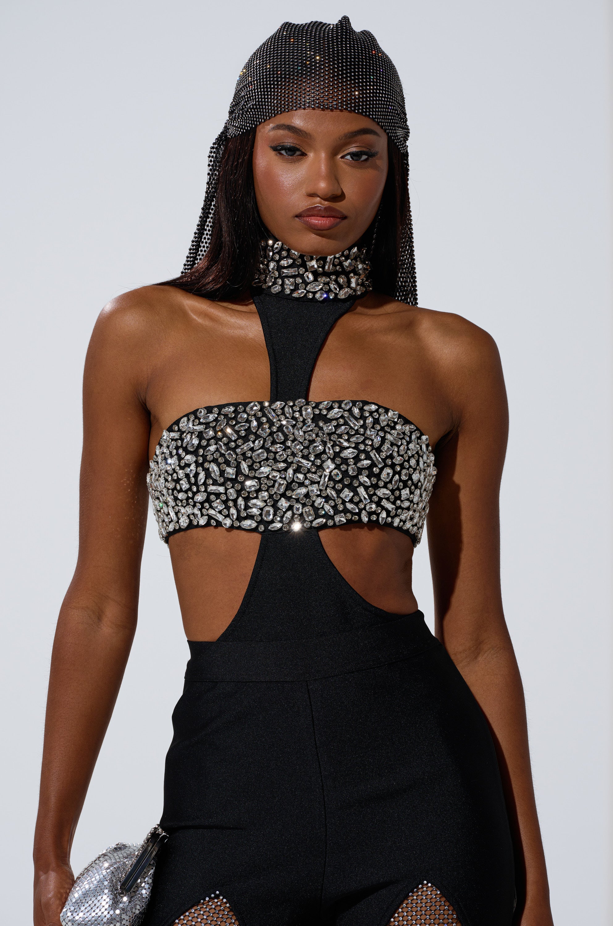 A woman stands confidently against a plain background, wearing THE BEST HAS YET TO COME RHINESTONE BODYSUIT—a sparkling piece with silver embellishments—paired with a beaded headpiece and a matching silver purse.