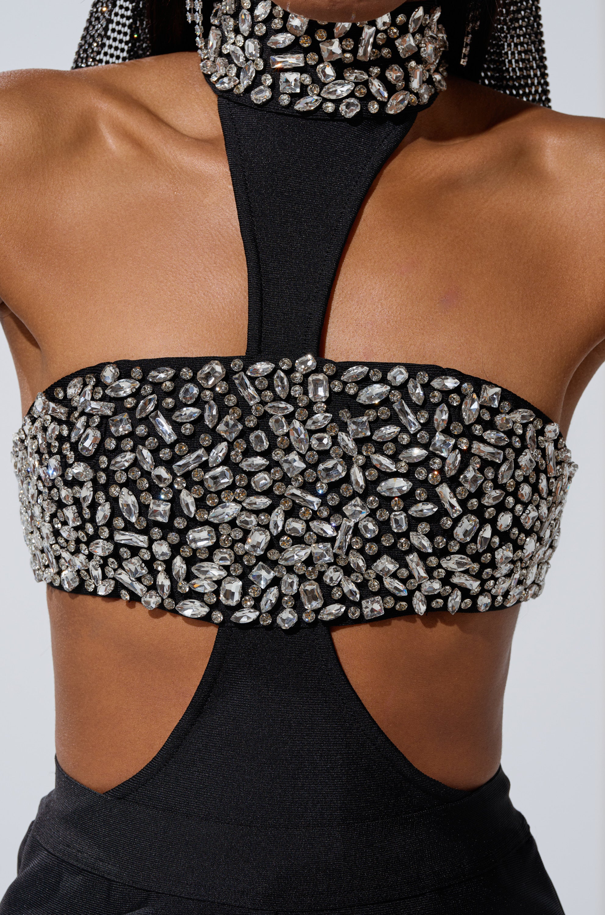 A close-up of a person wearing THE BEST HAS YET TO COME RHINESTONE BODYSUIT, a black, stretchy bandage halter with sparkling rhinestone clusters, attached rhinestone choker, and bare shoulders and upper back.