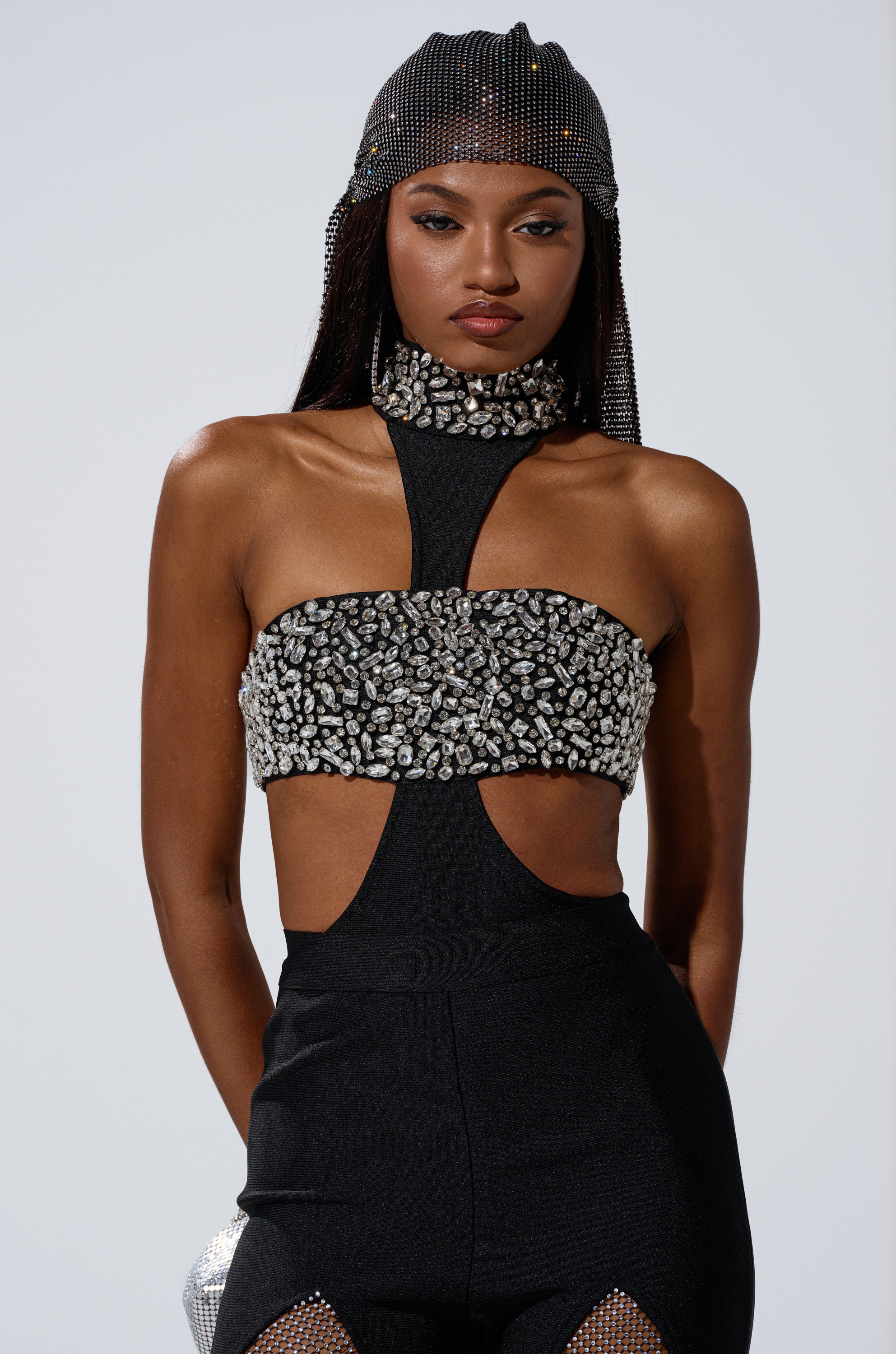 A woman models THE BEST HAS YET TO COME RHINESTONE BODYSUIT, featuring dazzling rhinestones and bold cut-outs, paired with a beaded headpiece, as she stands against a plain background.