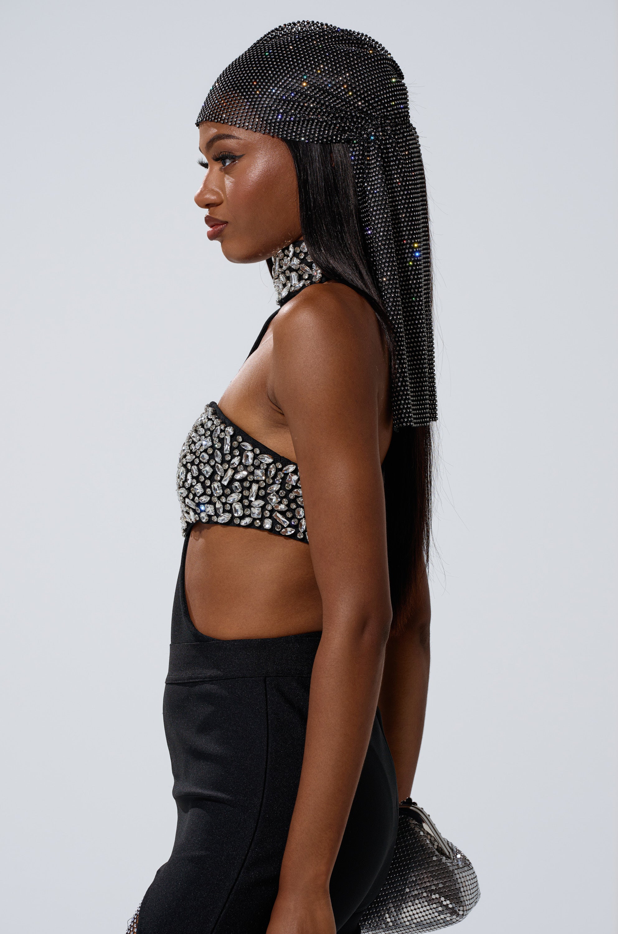 A woman in profile wears THE BEST HAS YET TO COME RHINESTONE BODYSUIT with a cut-out front, a jeweled headscarf, and high-waisted black pants against a plain light gray background.