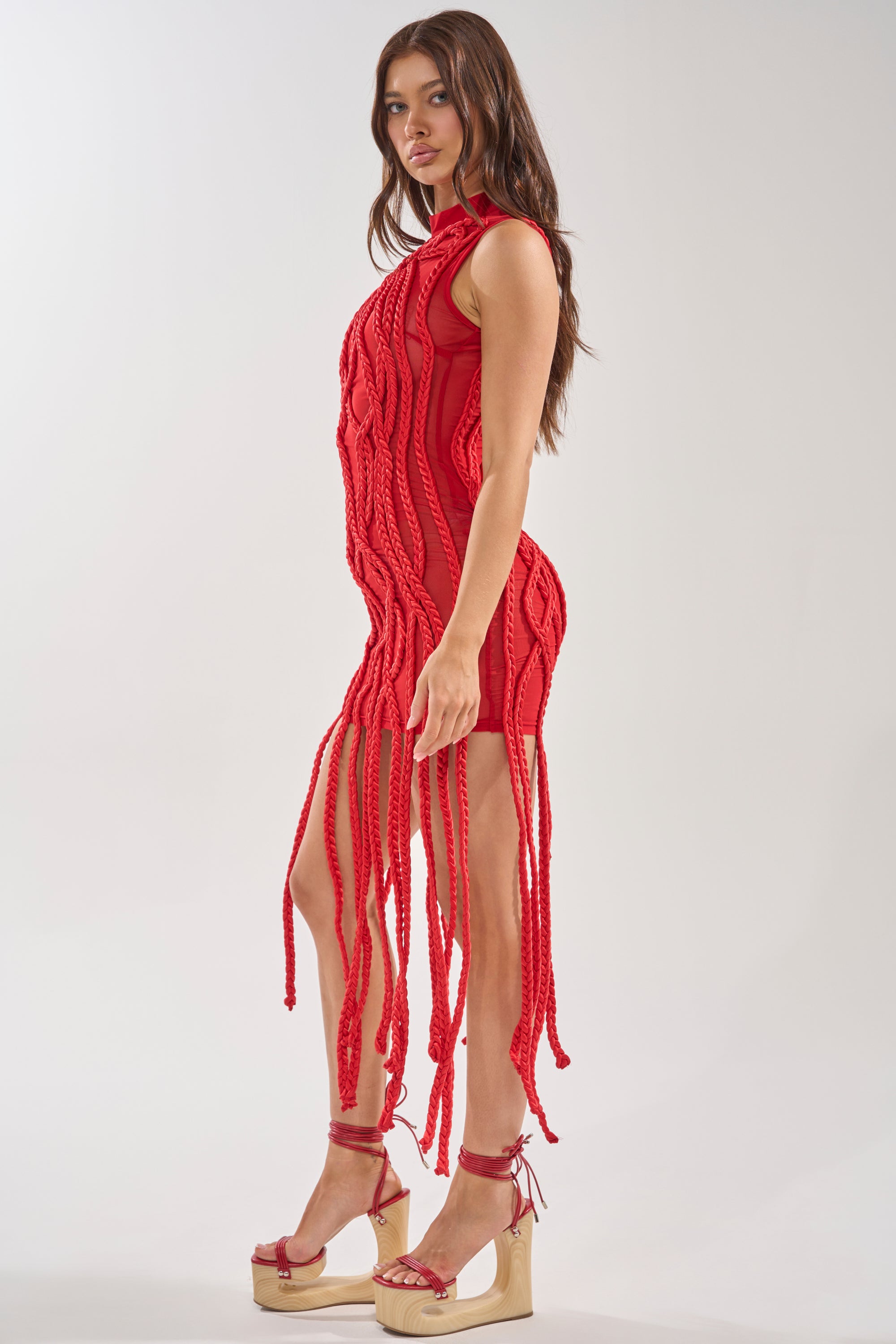 A woman models the STUCK ON YOU BRAIDED MINI DRESS IN RED, featuring bold braided details and long fringe, styled with matching red platform heels. She stands sideways against a light background, looking toward the camera. 2