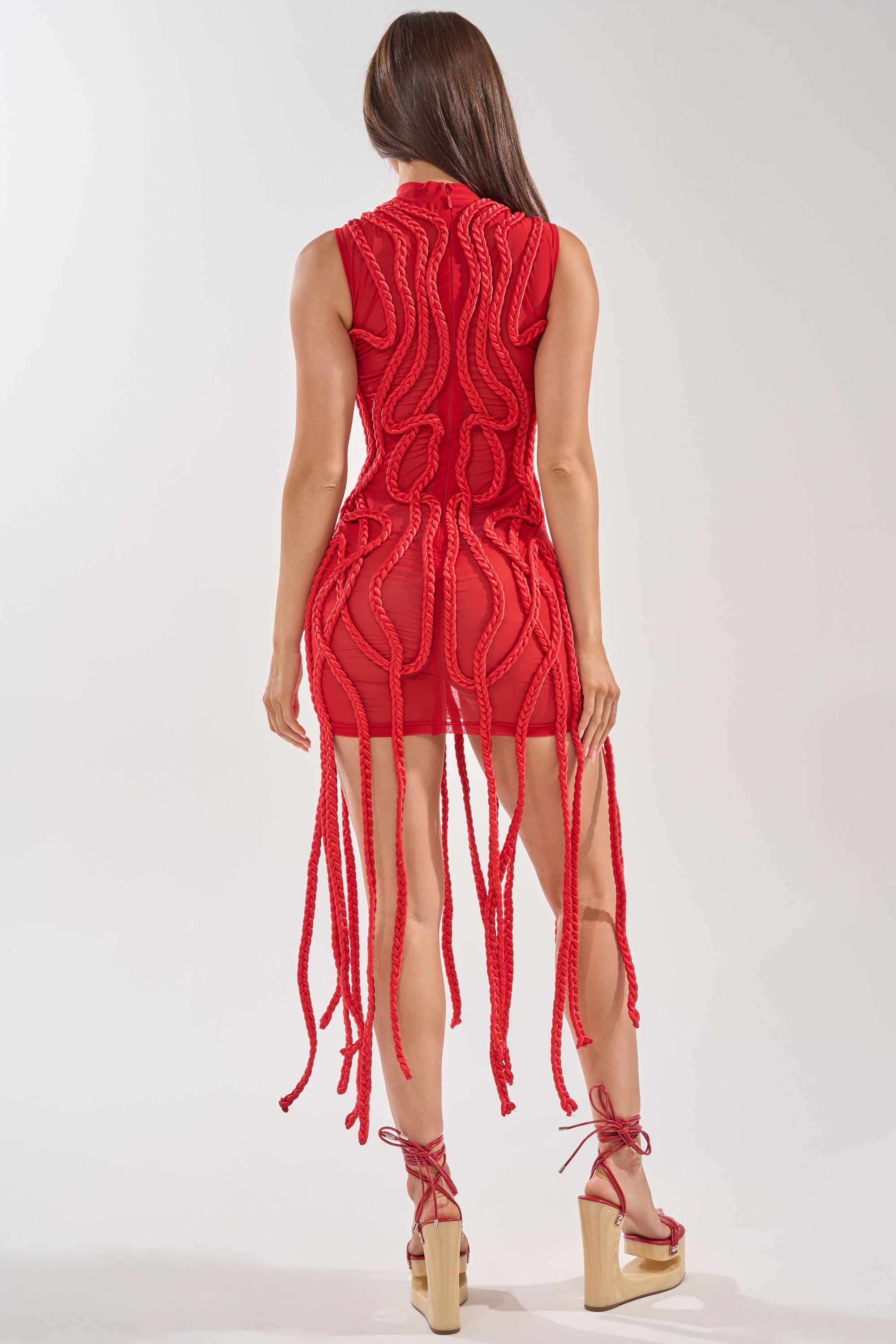 A woman with long brown hair wears the STUCK ON YOU BRAIDED MINI DRESS IN RED, a sleeveless, form-fitting mini dress featuring thick braided details down the back. She pairs it with red lace-up wedge sandals against a plain light background. 3
