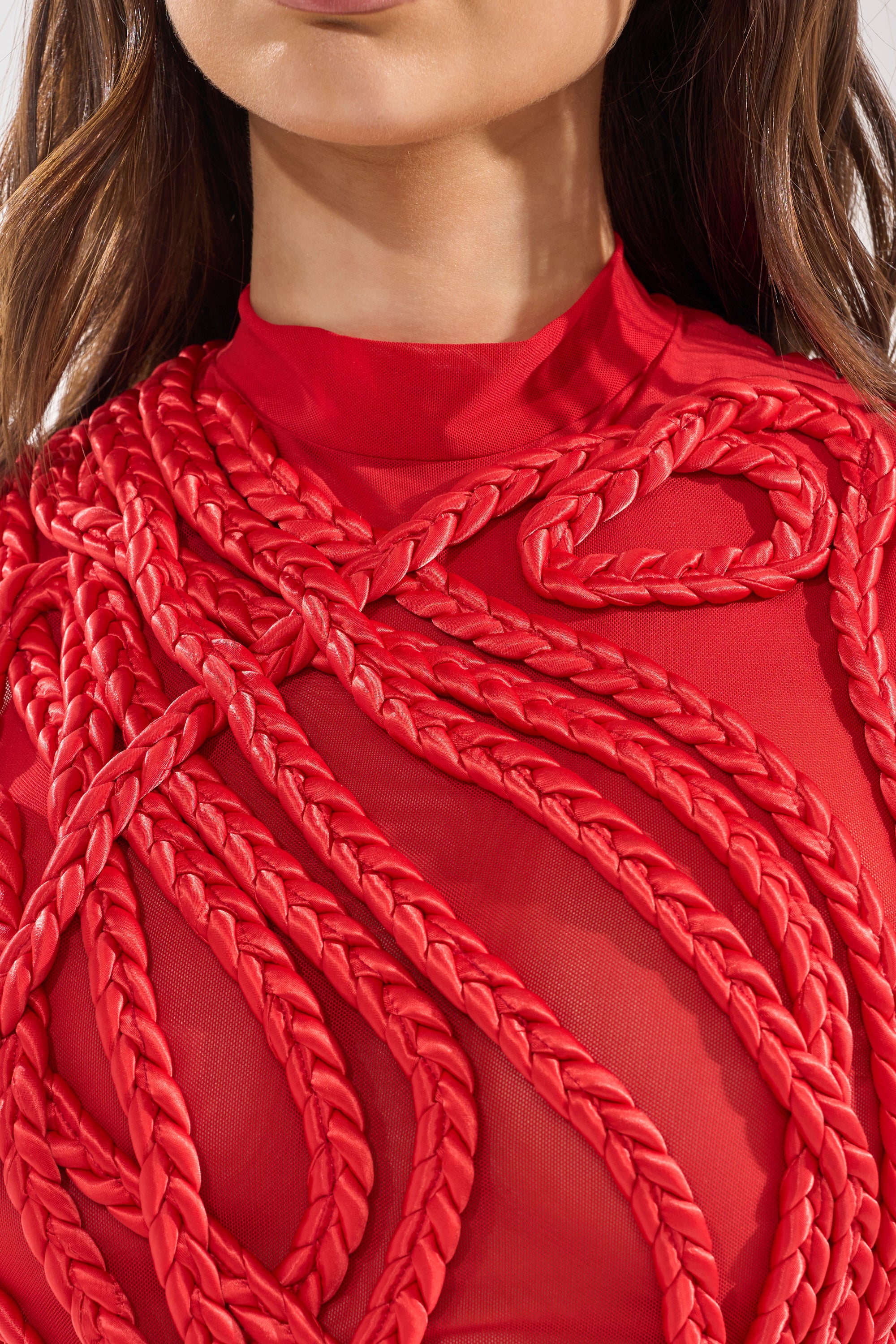 A woman models the STUCK ON YOU BRAIDED MINI DRESS IN RED, featuring sheer fabric and bold, thick braided red ropes in striking patterns across her chest and shoulders. Her lower face and torso are shown, emphasizing the dress’s unique braided details. 4
