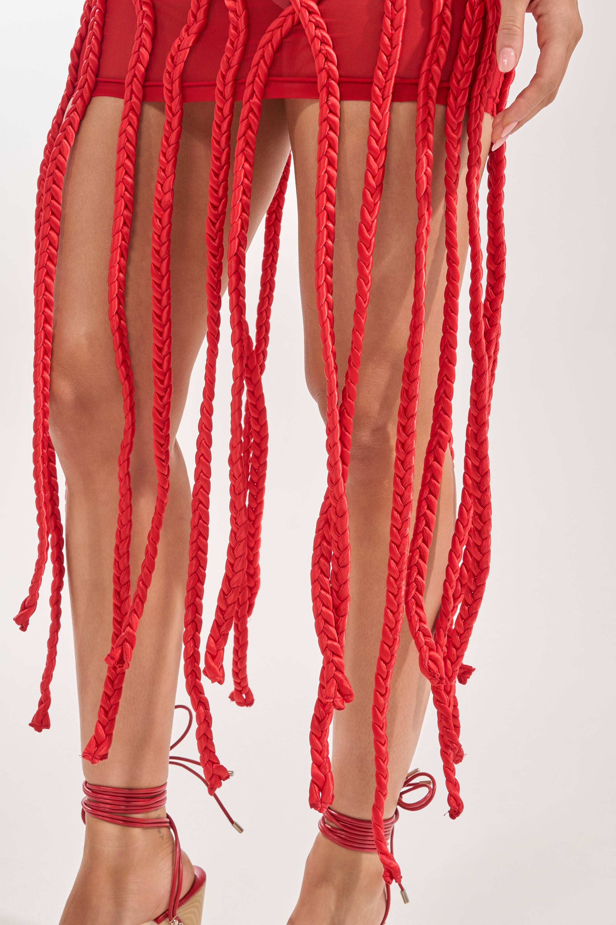 A person wears the STUCK ON YOU BRAIDED MINI DRESS IN RED, featuring long, thick braided red tassels, paired with red strappy sandals against a plain light-colored background. 5