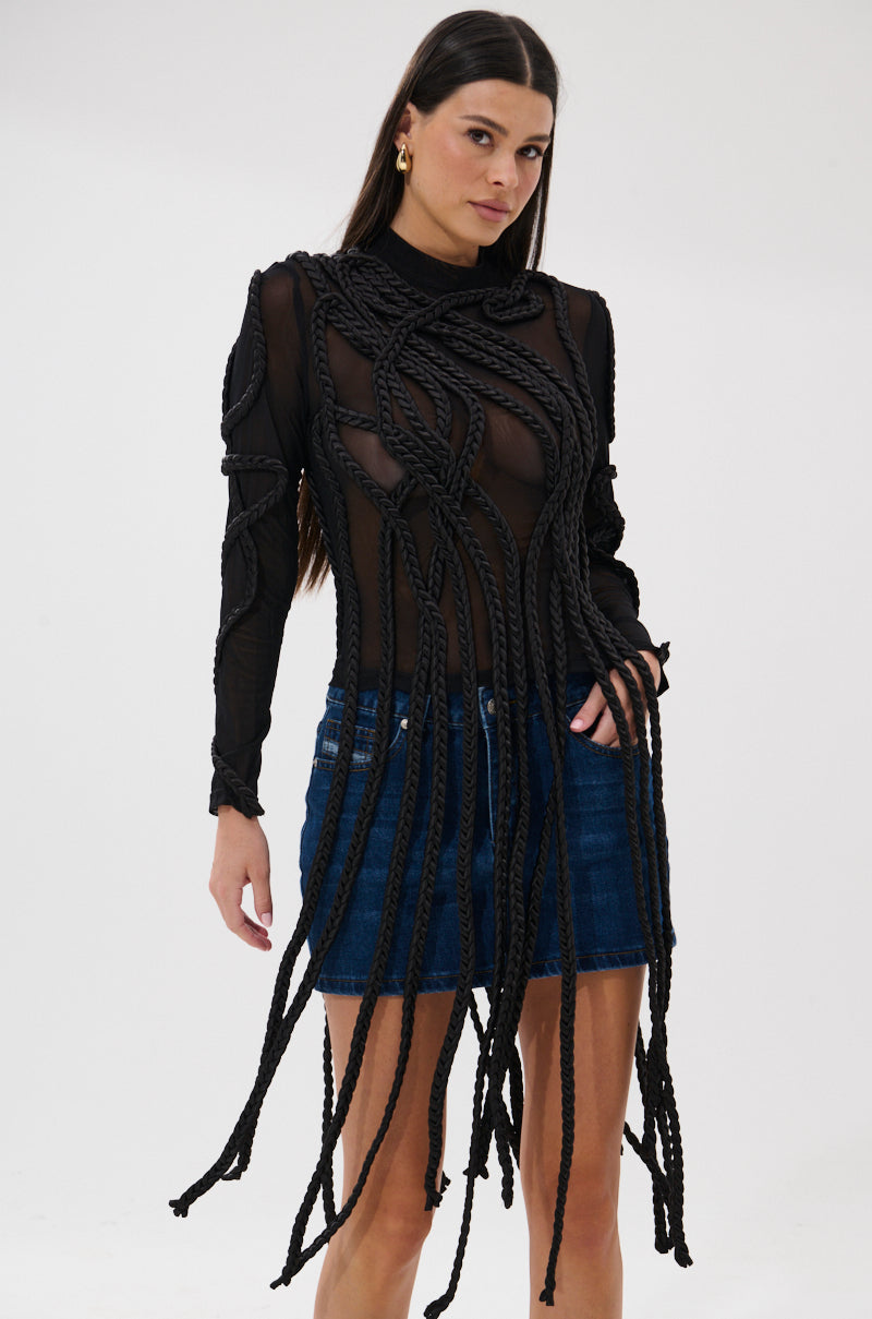 A woman models the ROPE YOU IN LONG SLEEVE MESH TOP—sheer black mesh with braided rope details—paired with a blue denim skirt, set against a plain white background.