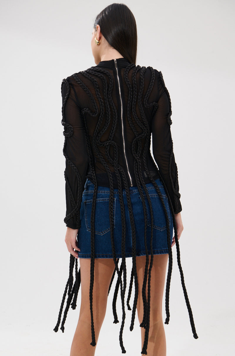 A person faces away, wearing the ROPE YOU IN LONG SLEEVE MESH TOP featuring braided rope detail down the back, paired with a denim mini skirt. The background is plain and light-colored.