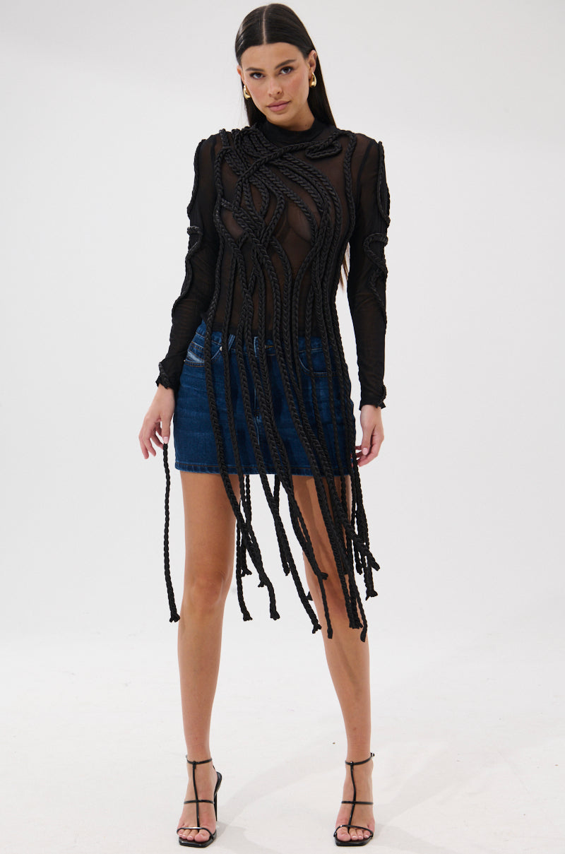 A woman models the ROPE YOU IN LONG SLEEVE MESH TOP featuring intricate braided rope detail, styled with a blue denim mini skirt and black strappy heels, posing confidently against a plain white background.