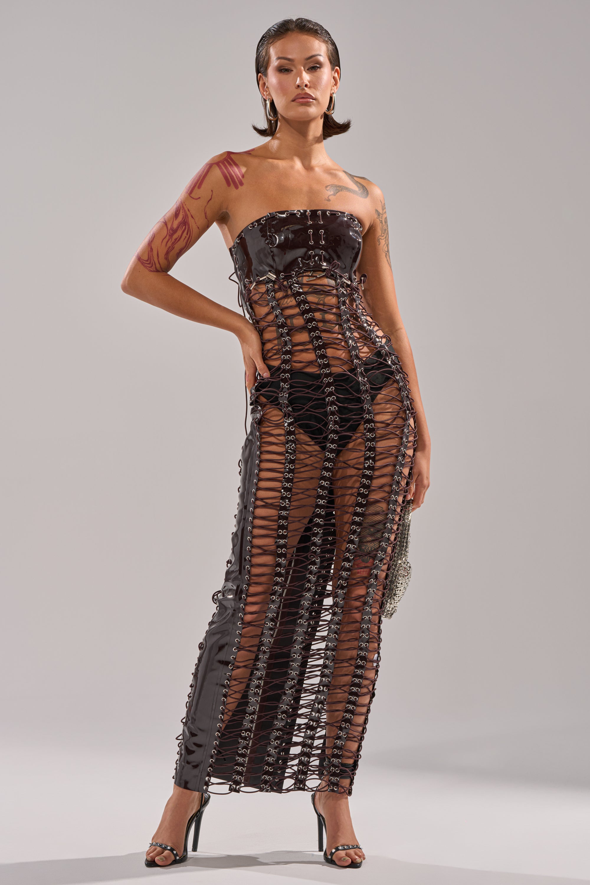 A model poses in the ONE OF ONE LACE UP MAXI DRESS—a strapless, glossy faux patent leather gown with sheer sections that reveal undergarments. She pairs it with black heels, a small bag, and wears her hair slicked back. 3