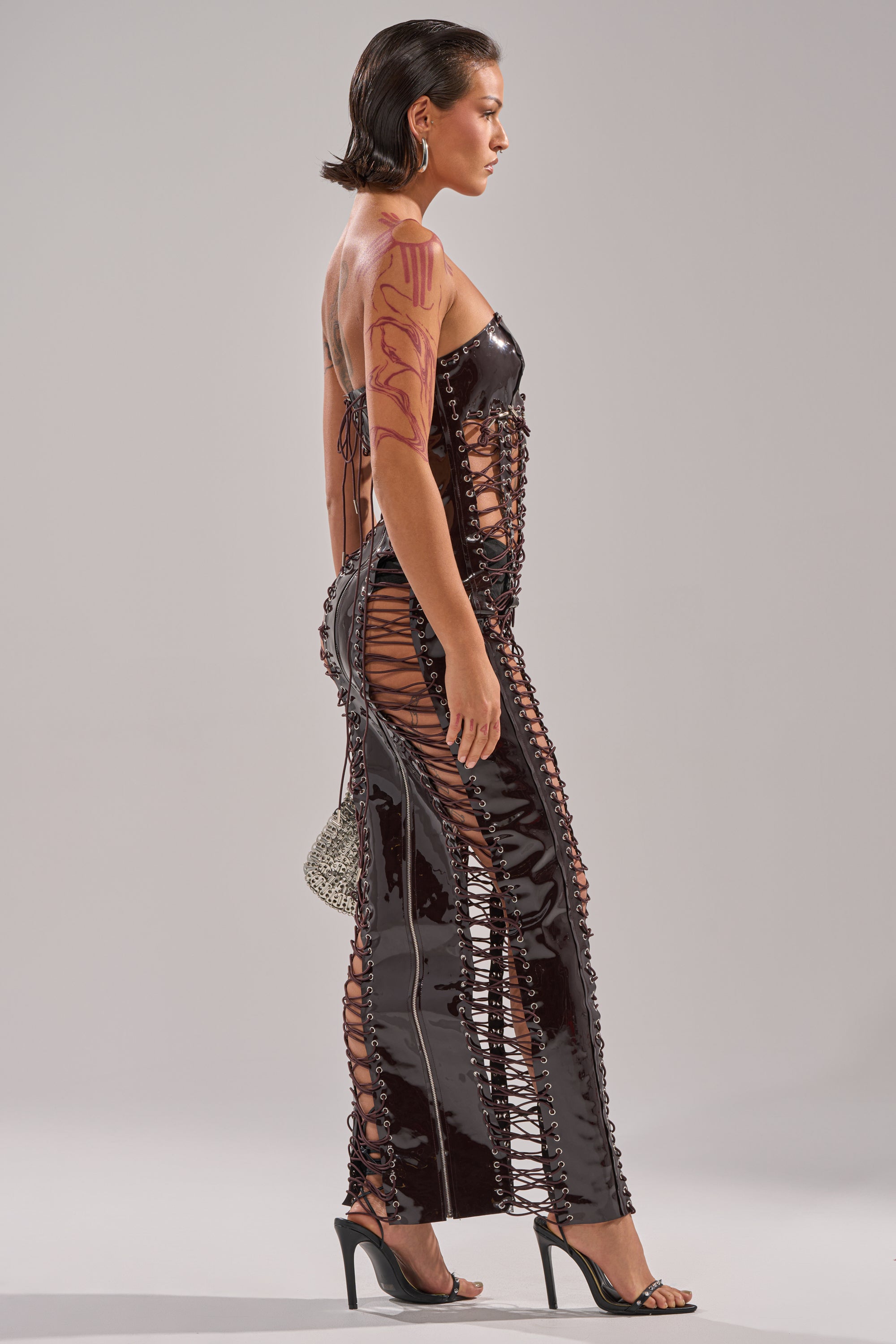 A woman in profile wears the ONE OF ONE LACE UP MAXI DRESS, a black faux patent leather cutout maxi with lace-up detailing, styled with high heels, a sparkling mini handbag, short wet-look hair, and bold upper-arm body art. 4