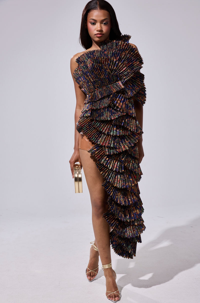 A woman poses confidently in the LUST FOR LIFE ASYMMETRICAL BROCADE TOP, featuring a one-shoulder neckline and ruffled, iridescent layers. She holds a gold clutch and wears strappy heels against a neutral background.