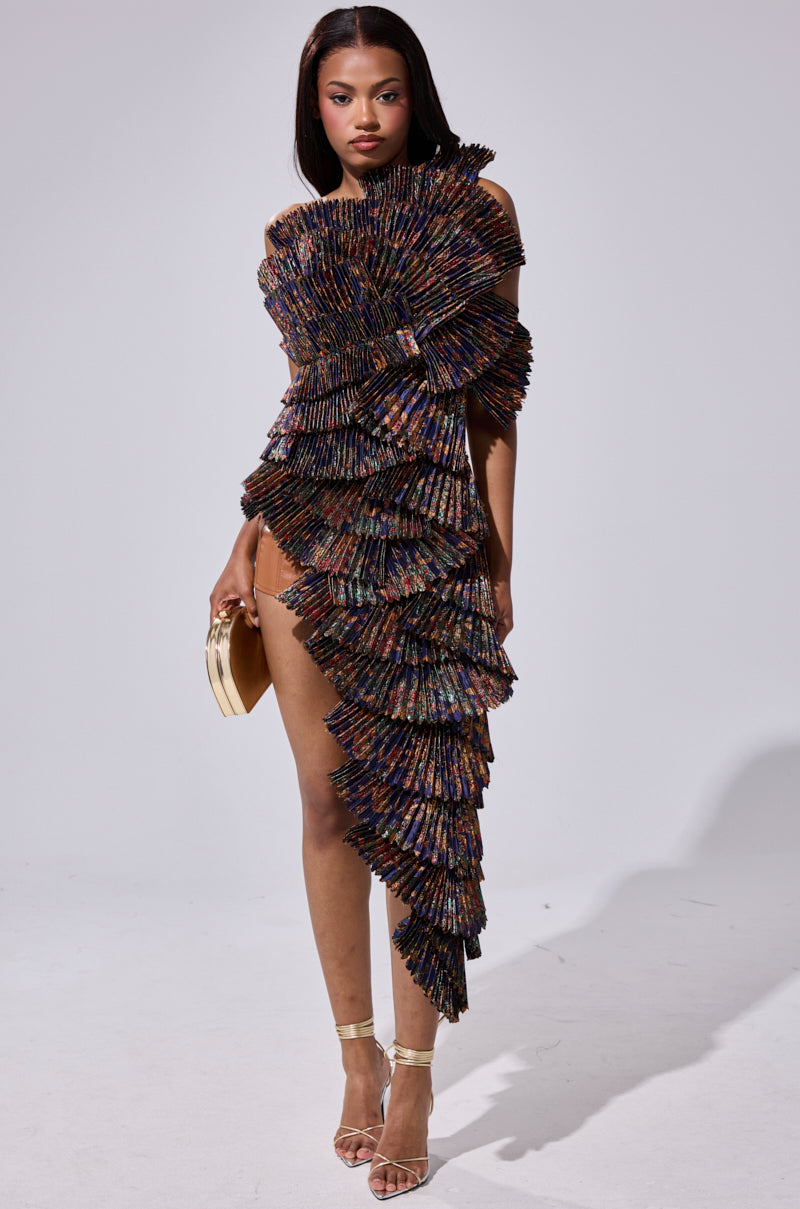 A woman stands confidently in the LUST FOR LIFE ASYMMETRICAL BROCADE TOP, featuring layered, fan-like pleated panels in multicolored hues, paired with a gold clutch and heels, against a plain light background.