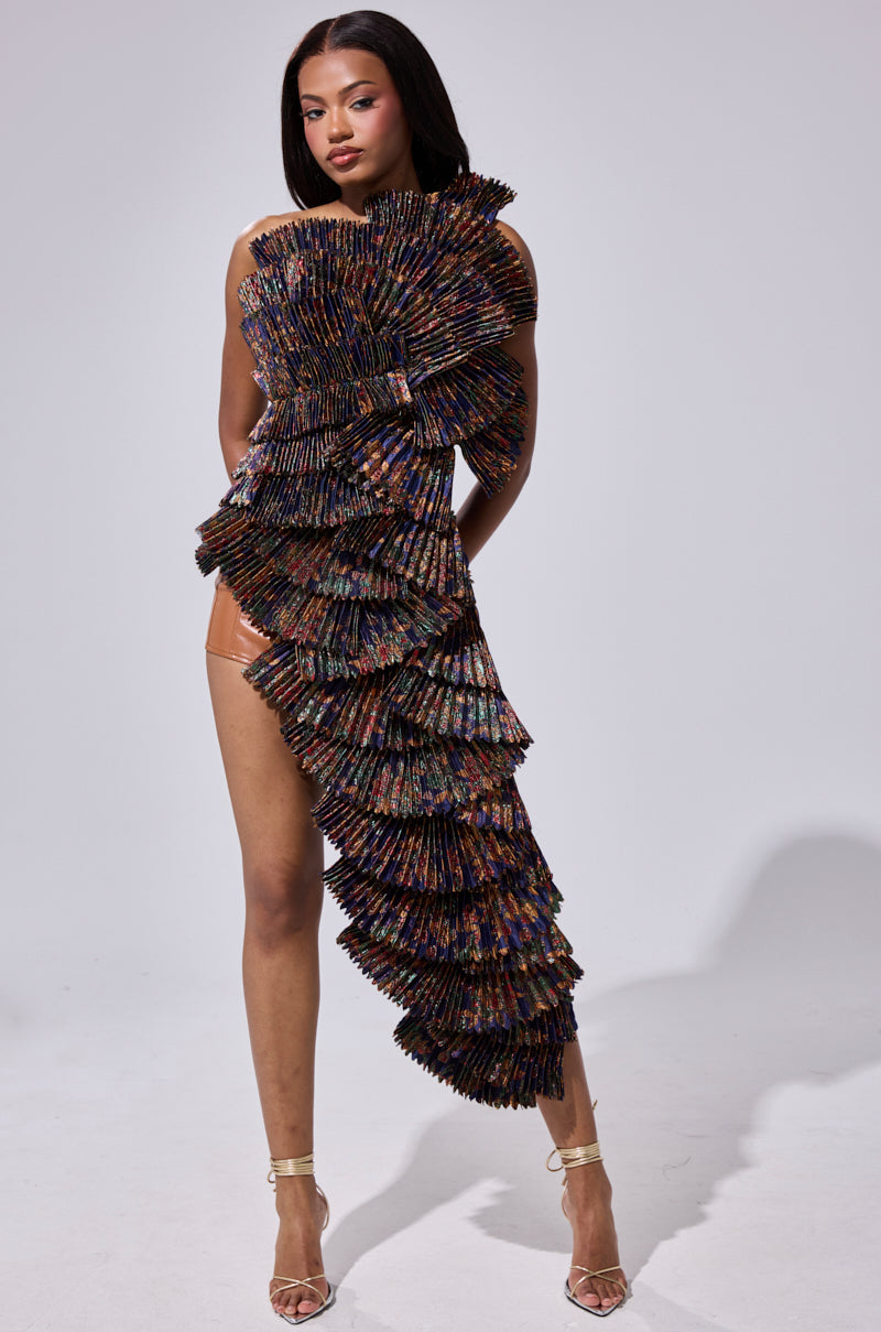 A woman stands confidently in the LUST FOR LIFE ASYMMETRICAL BROCADE TOP with pleated, multicolored panels and a dramatic one-shoulder neckline, paired with strappy gold heels against a plain light background.