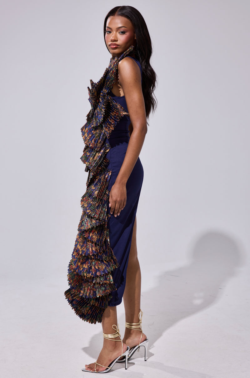 A woman stands sideways wearing the LUST FOR LIFE ASYMMETRICAL BROCADE TOP, featuring bold multicolored ruffles and pleated front panels. She has long, wavy dark hair and pairs the look with strappy high-heeled sandals.
