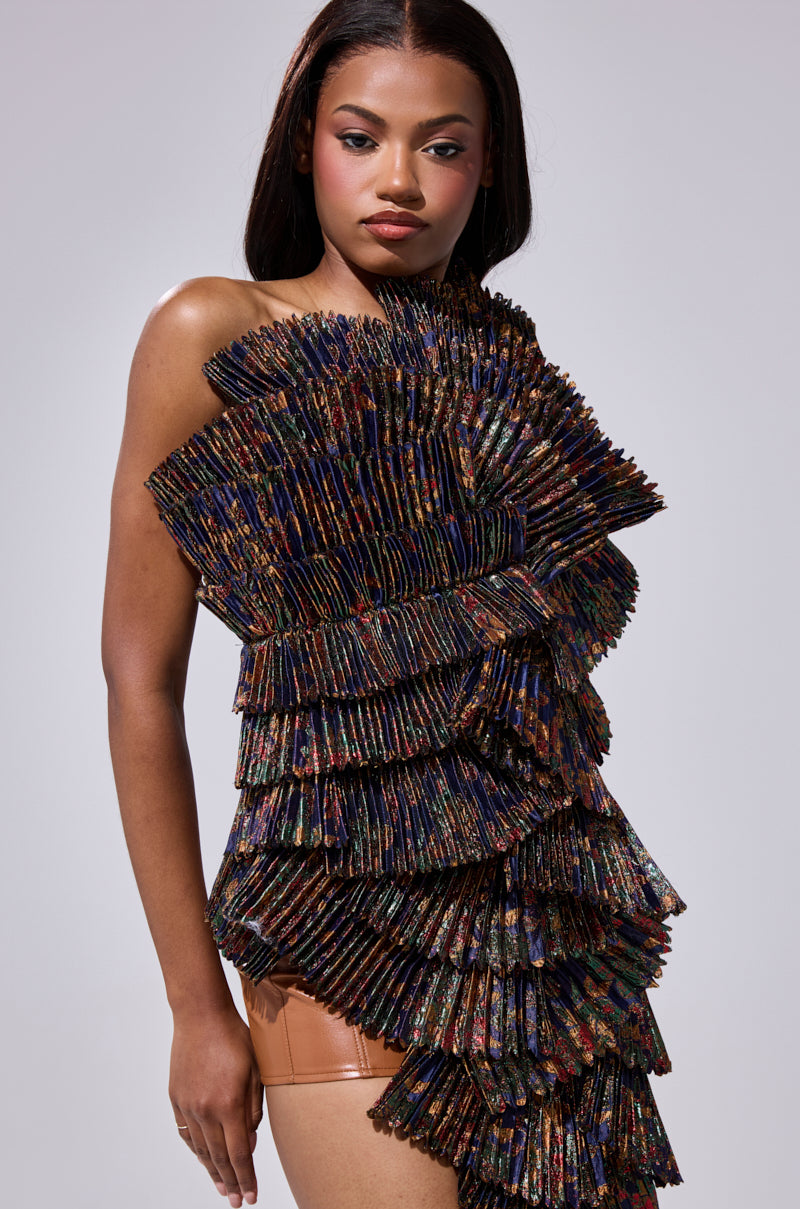 A woman with straight dark hair and natural makeup poses confidently in the LUST FOR LIFE ASYMMETRICAL BROCADE TOP, featuring a sculptural one-shoulder neckline, layered pleated panels, and multicolored metallic fabric with a fan-like texture.