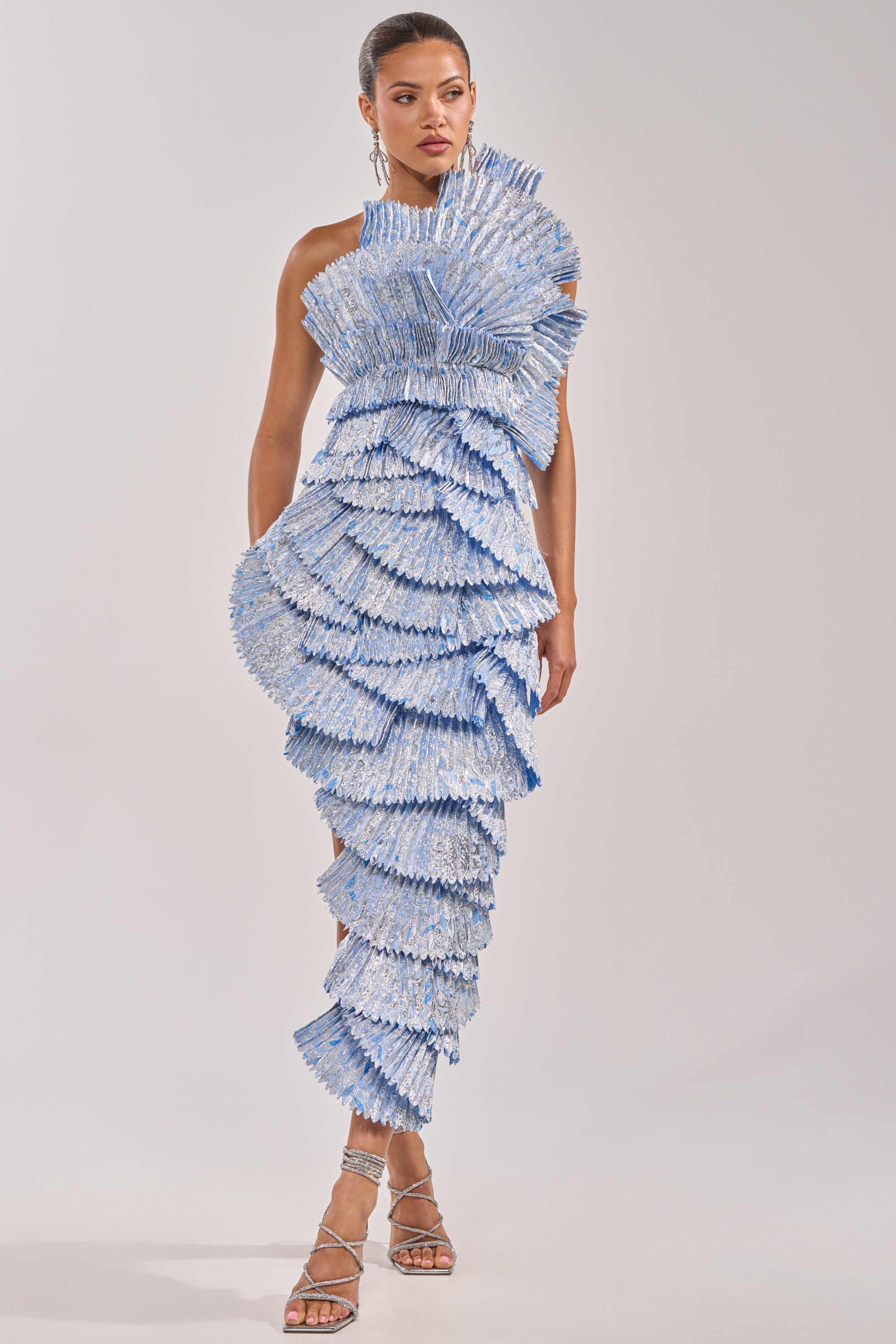A woman models the LUST FOR LIFE Asymmetrical Brocade Midi Dress in Blue, featuring sculptural ruffles, fan-like textures, a one-shoulder neckline, and strappy heels against a plain light background. 0