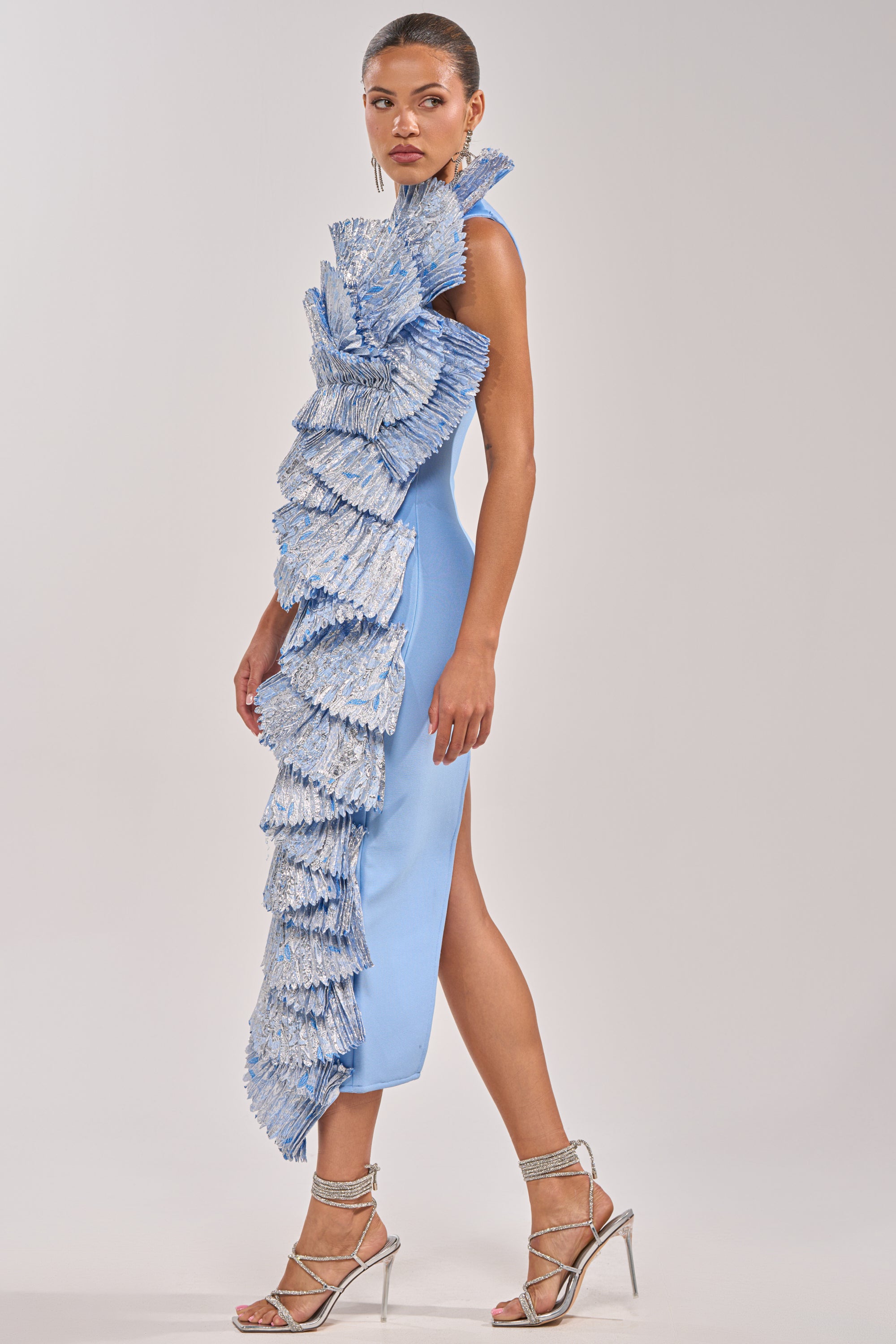 A woman stands in profile wearing the LUST FOR LIFE ASYMMETRICAL BROCADE MIDI DRESS IN BLUE, featuring dramatic ruffled, fan-like embellishments down the front. Her hair is slicked back and she pairs the look with silver strappy high heels. 2