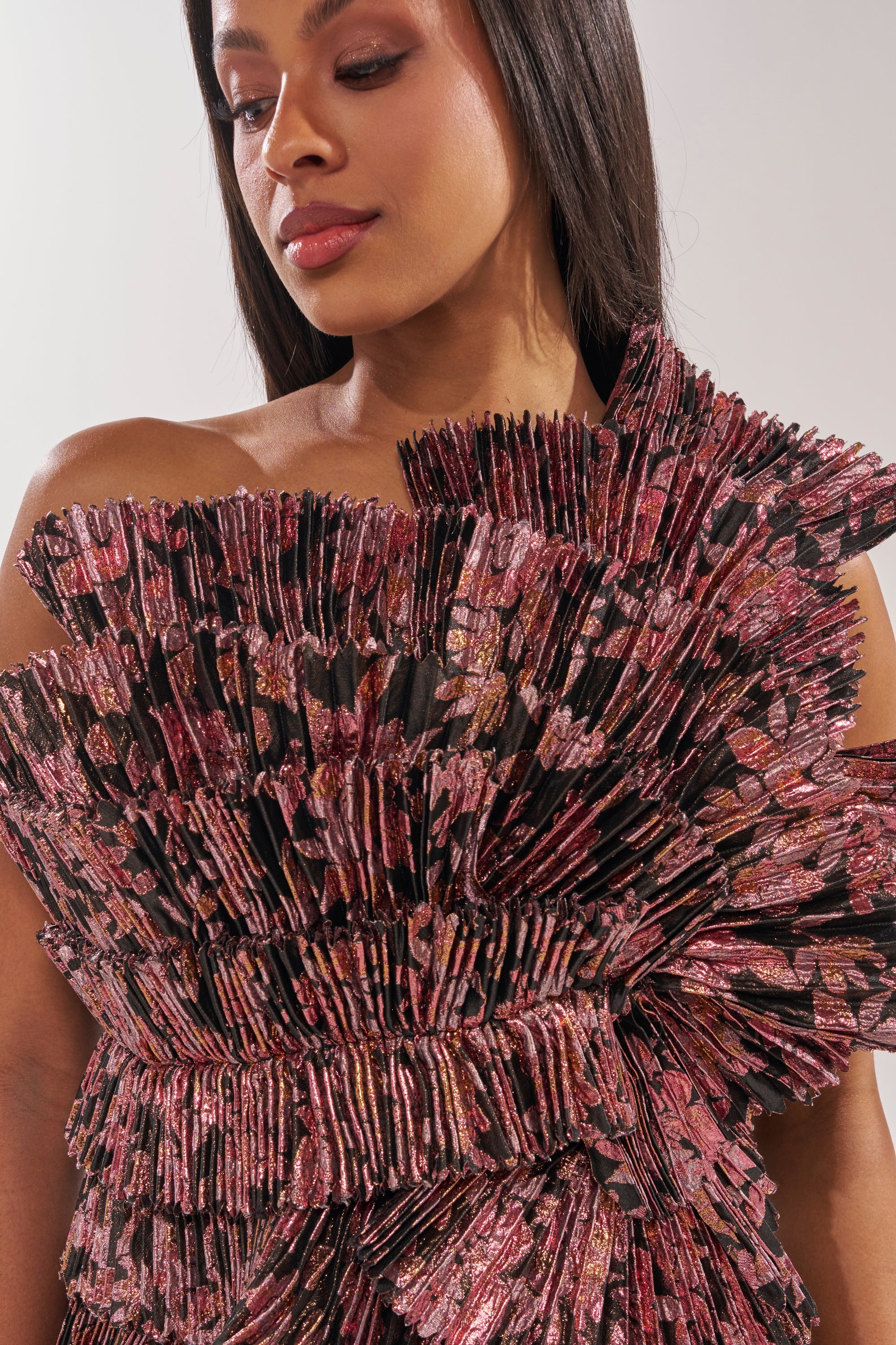 A woman with straight dark hair wears the RESTLESS NIGHTS ASYMMETRICAL BROCADE TOP, featuring pleated texture and shimmering pink, black, and gold ruffled layers in a one-shoulder silhouette that highlights its asymmetrical design. 4