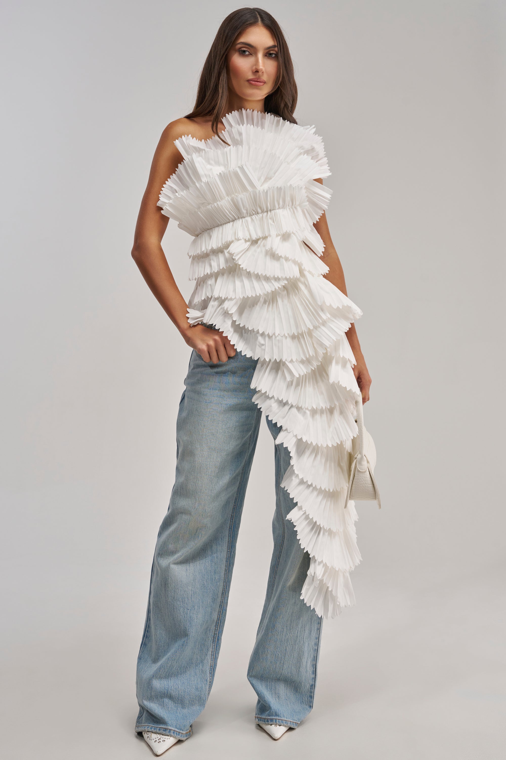 A woman poses in wide-leg light blue jeans and the LUXURIOUS ASYMMETRICAL TOP IN WHITE, featuring dramatic pleats and layered ruffles. She accessorizes with a small white handbag and pointed toe heels.