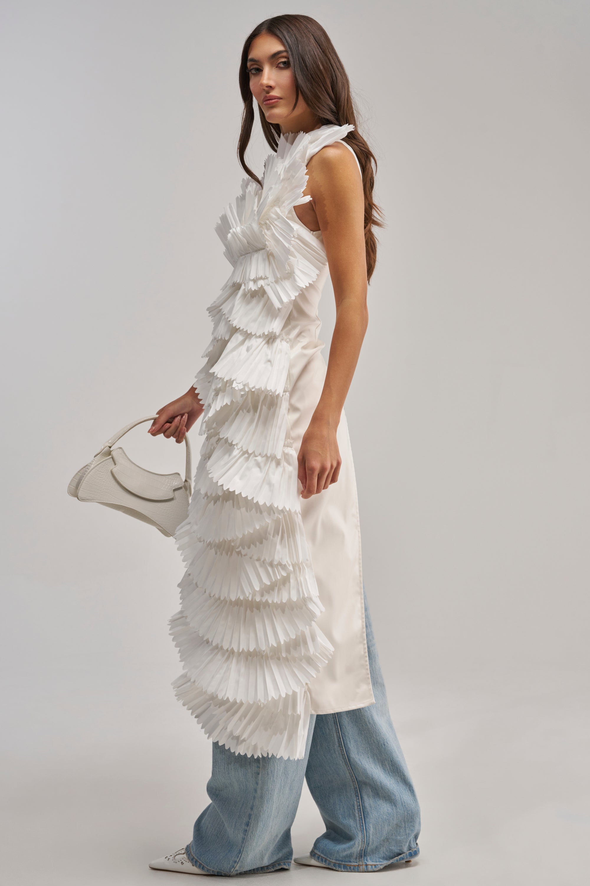 A woman poses against a plain background wearing the LUXURIOUS ASYMMETRICAL TOP IN WHITE, paired with light blue wide-leg jeans, white sneakers, and a white handbag.