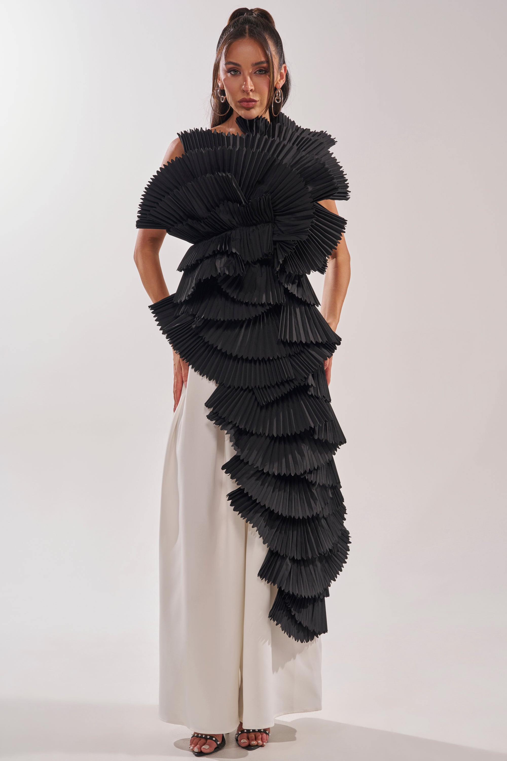 A woman stands against a plain background wearing the LUXURIOUS ASYMMETRICAL TOP IN BLACK with layered, pleated ruffle shoulders, paired with wide-legged white pants and black heels. 2