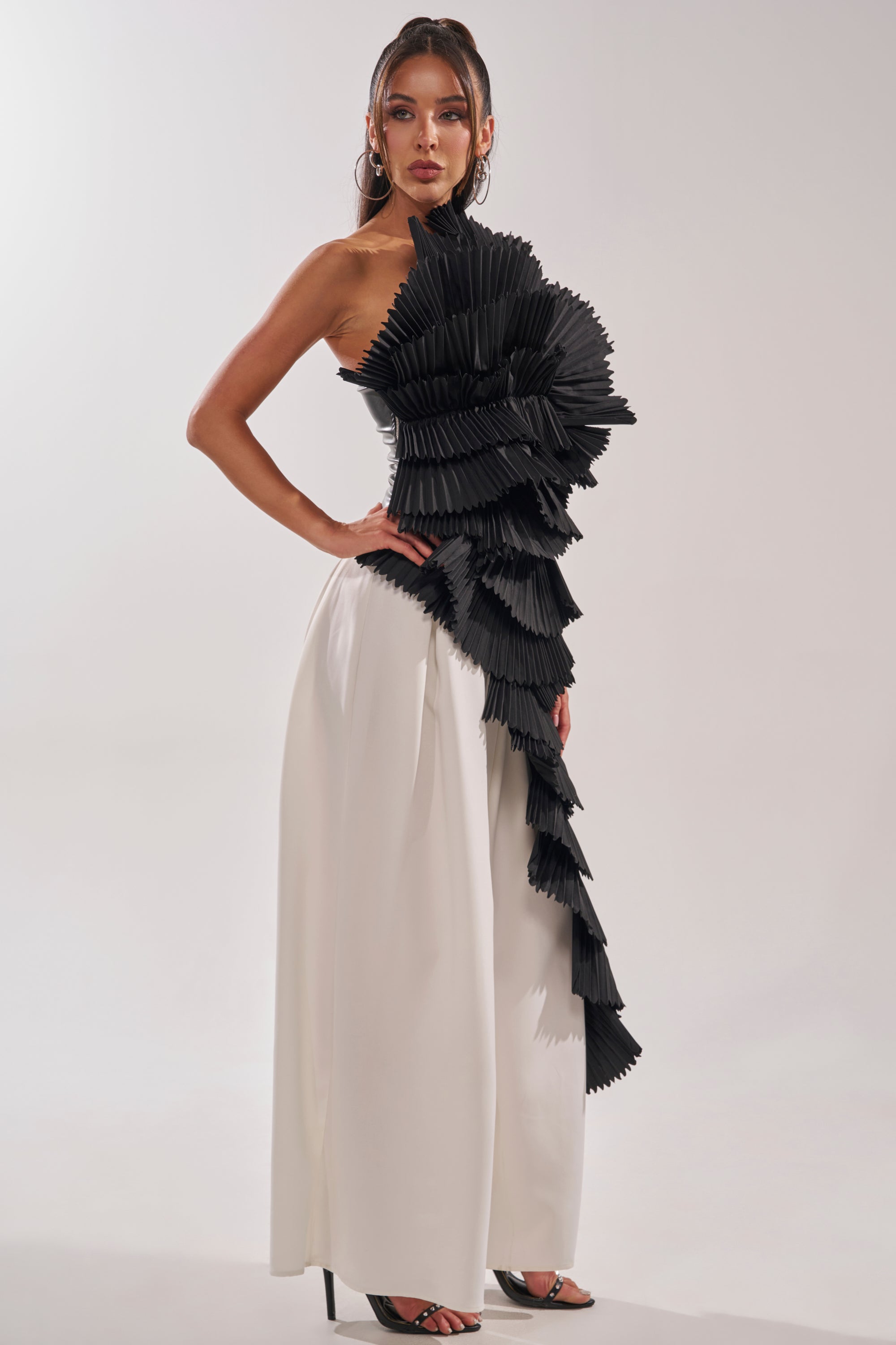 A woman poses in a modern look, featuring the LUXURIOUS ASYMMETRICAL TOP IN BLACK with wide-legged white pants, black open-toe heels, and hoop earrings. 4