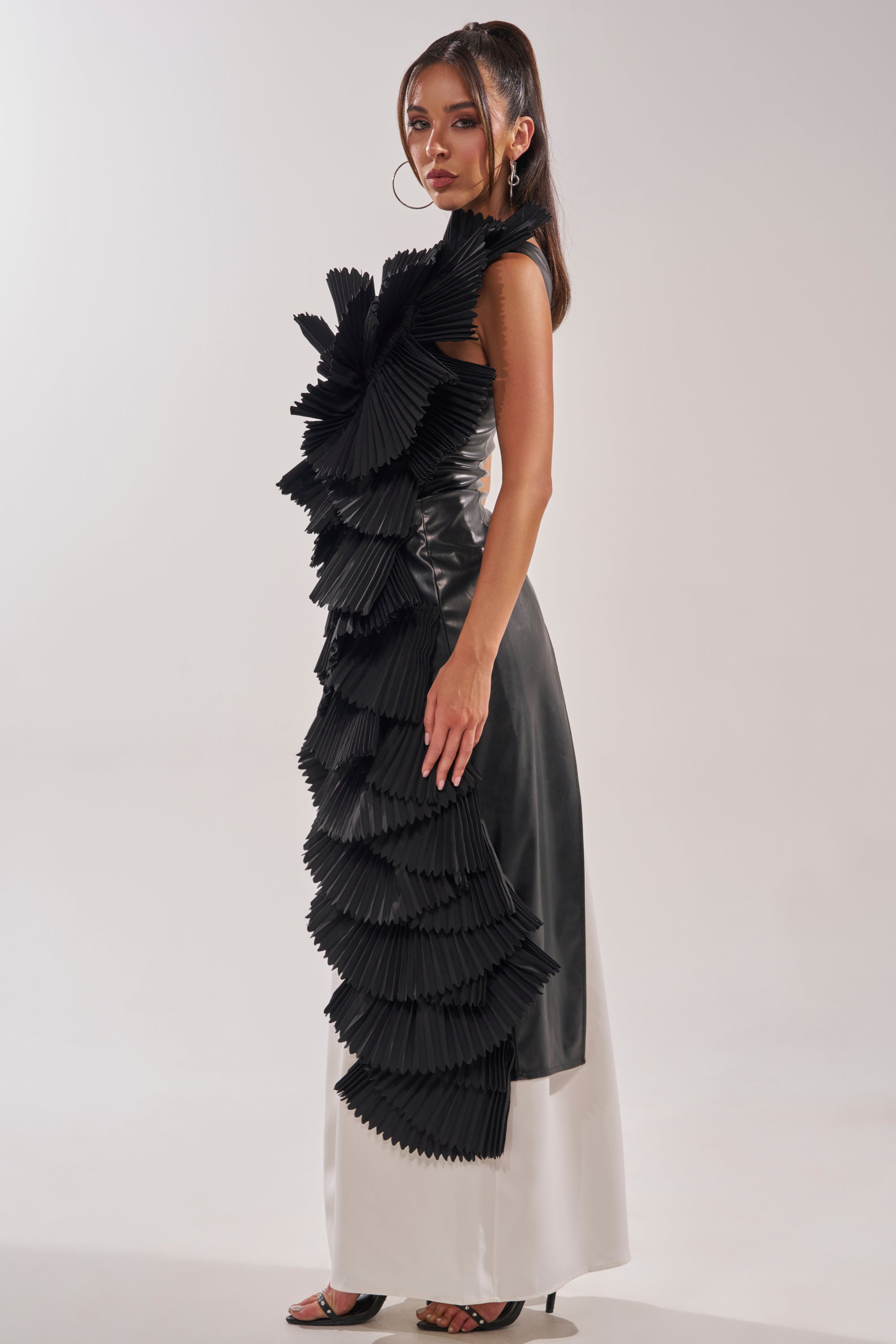A woman in the LUXURIOUS ASYMMETRICAL TOP IN BLACK, featuring a single-shoulder design with pleated sculptural ruffles and a long two-tone black and white skirt, stands sideways with long dark hair, hoop earrings, and strappy heels. 5