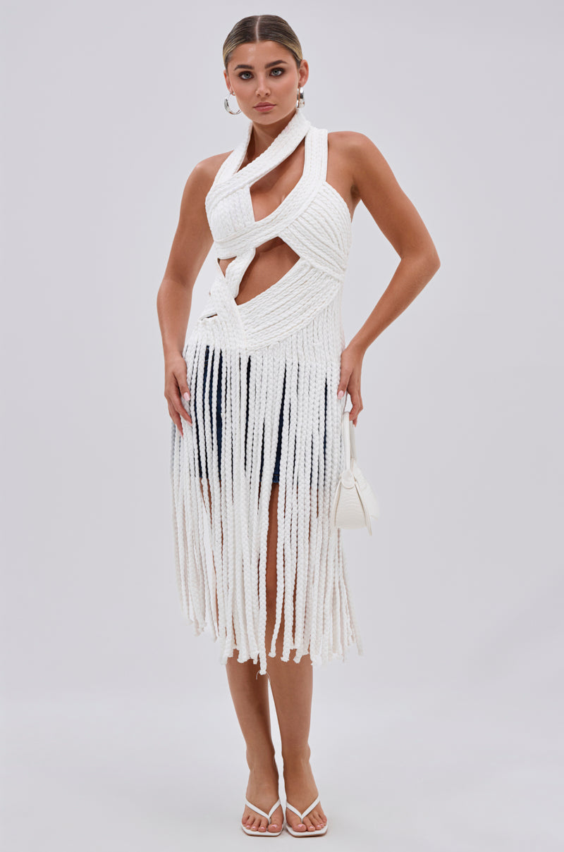 A woman stands against a light background wearing the CRAZY BUT THE SAME ROPE BLOUSE, a white intricately woven top with cutouts, fringe hem, and halter neckline. She accessorizes with a small white bag, white heels, and hoop earrings.