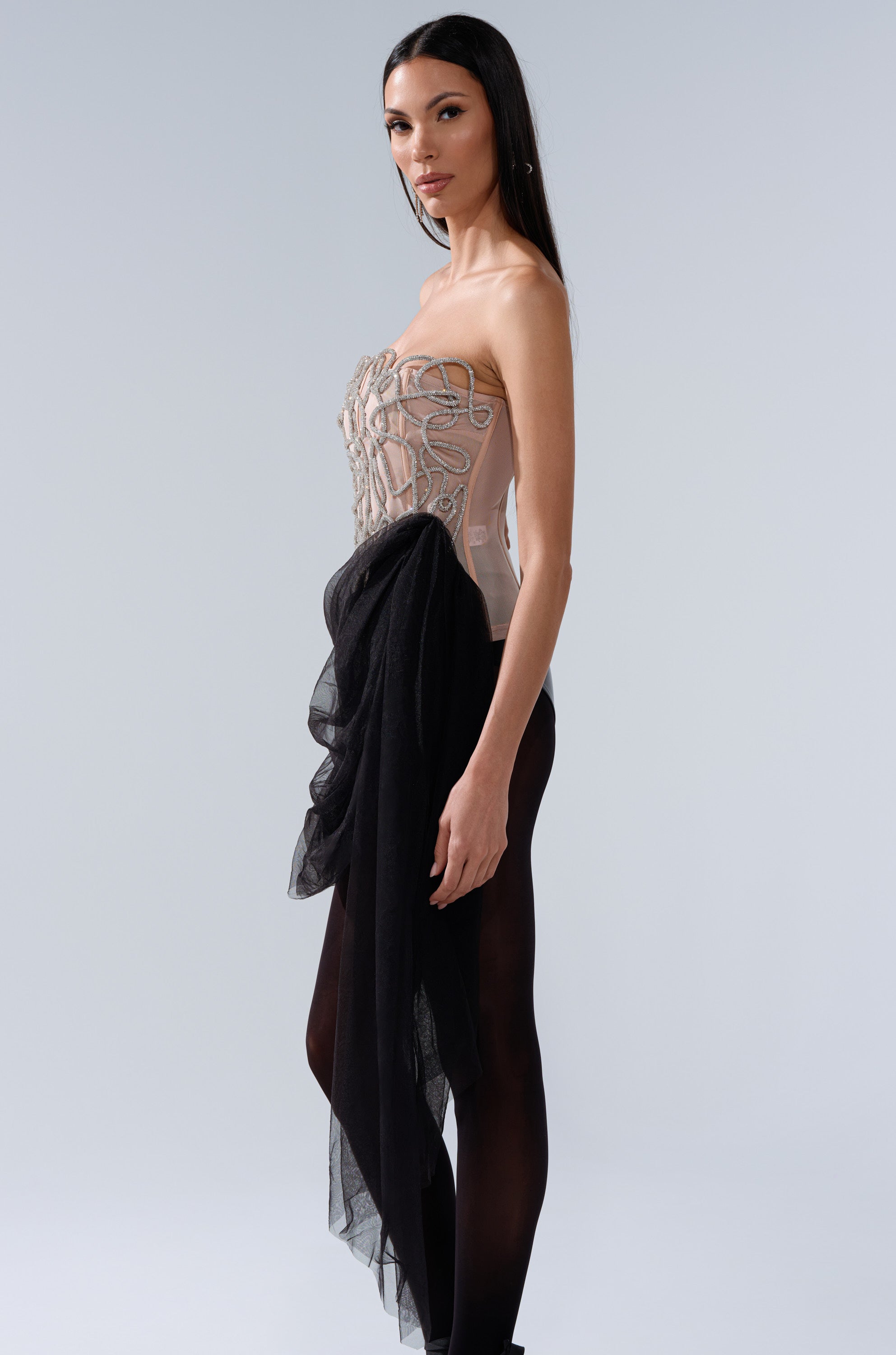 A woman with long dark hair stands sideways in the GIRL GO GET IT EMBELLISHED CORSET, styled with black sheer tights and a dramatic black tulle accent at her waist, against a light gray background.