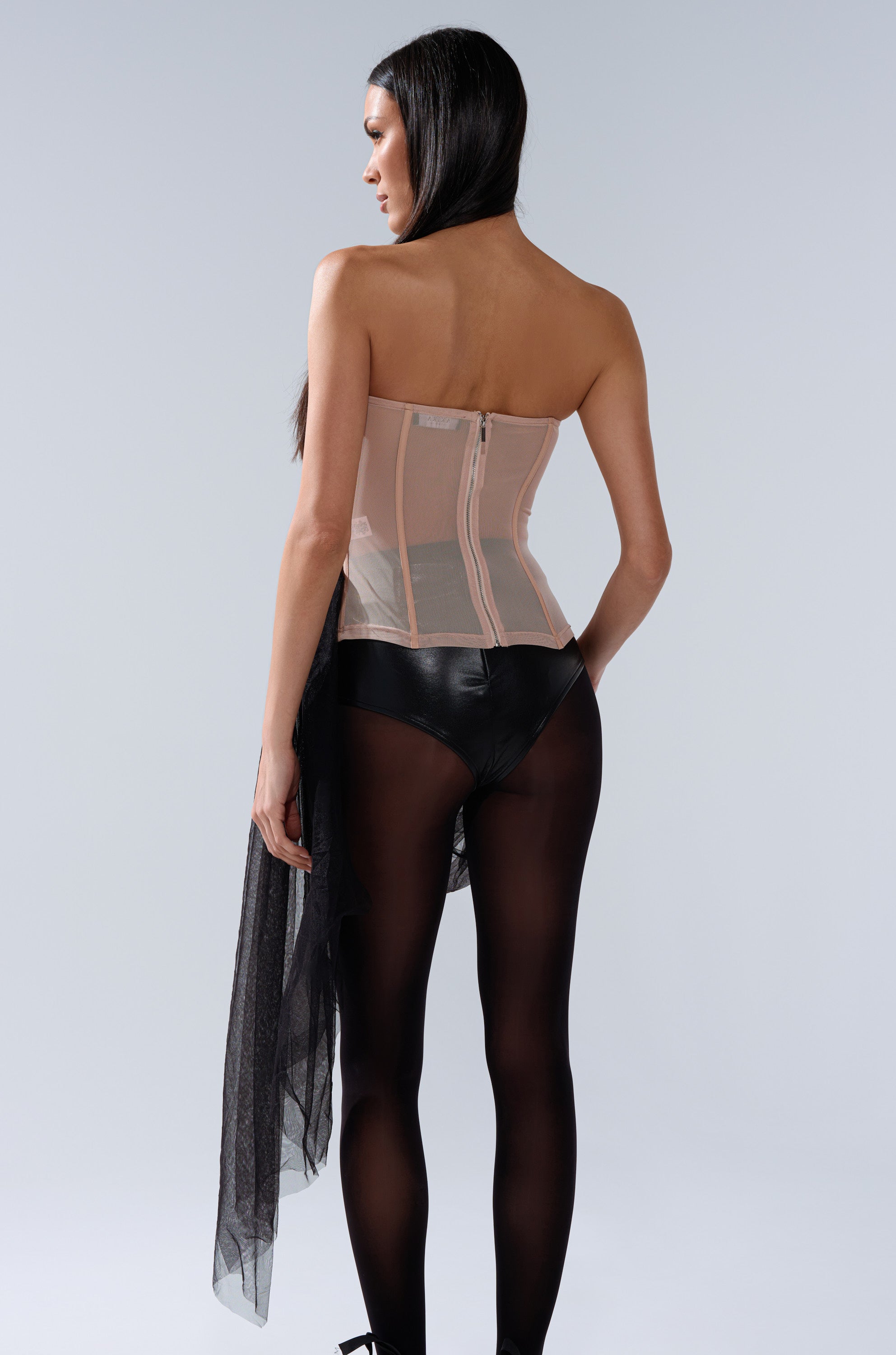 A woman with long dark hair stands facing away, wearing the GIRL GO GET IT EMBELLISHED CORSET, black high-waisted bottoms, and tights. She holds black tulle at her side against a light grey background, showcasing the corset's elegance.