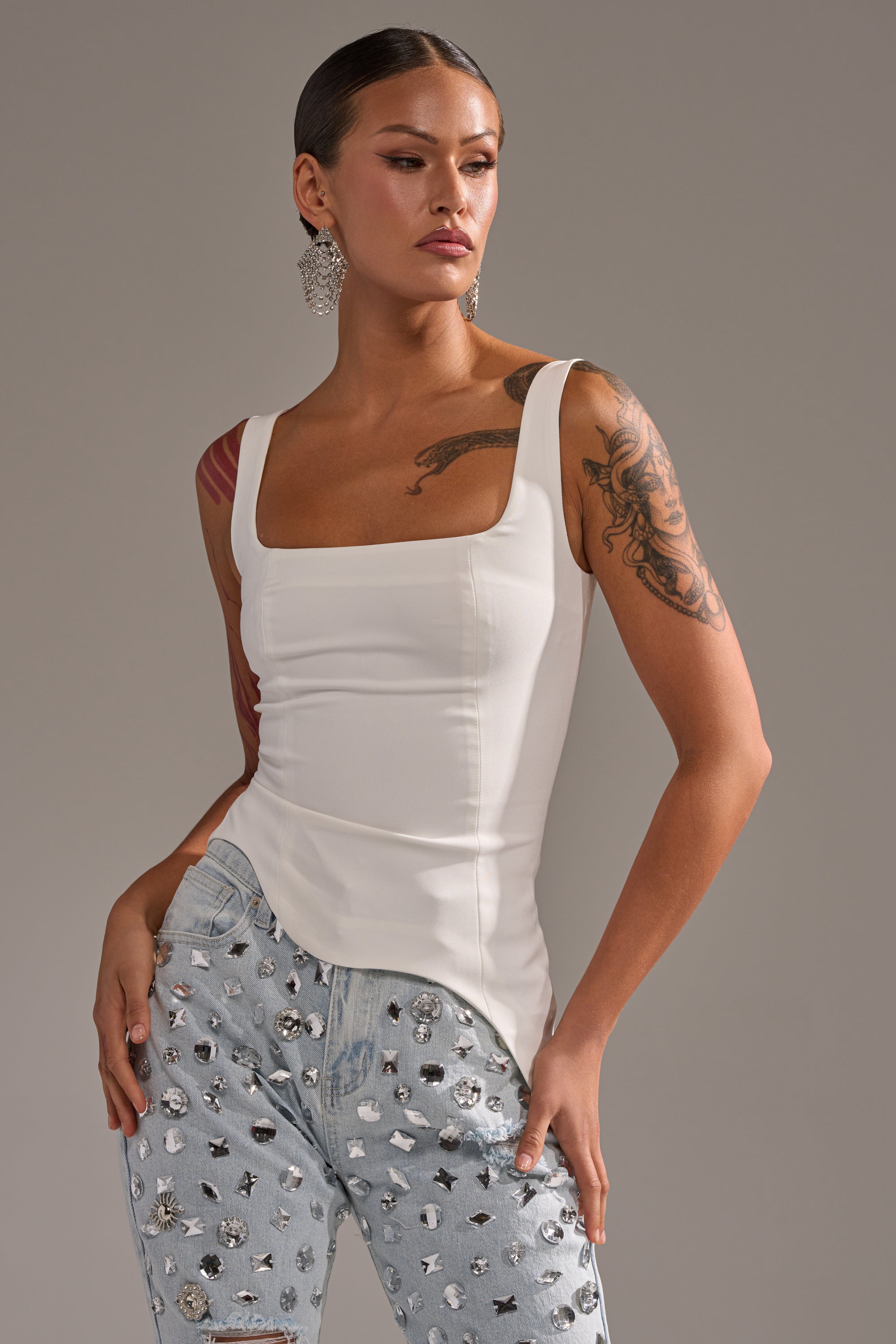 A woman with slicked-back hair and visible arm tattoos poses confidently in the PLAIN JANE SLEEVELESS BLOUSE, a white square-neck top. She completes her look with sparkly earrings and light blue rhinestone jeans. 0