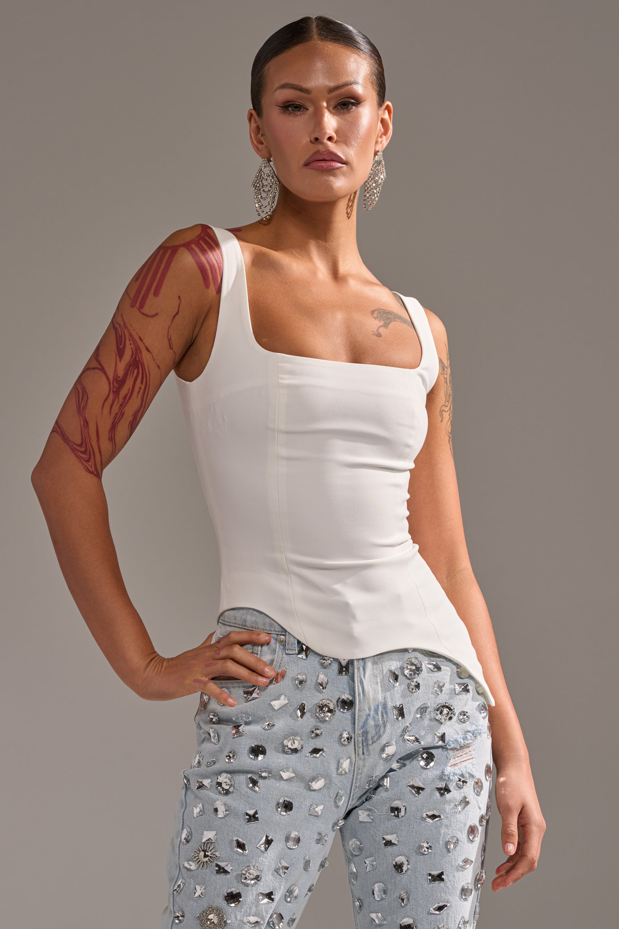 A person with long earrings and tattoos poses confidently in a fitted PLAIN JANE SLEEVELESS BLOUSE and light blue studded jeans against a plain background. 1