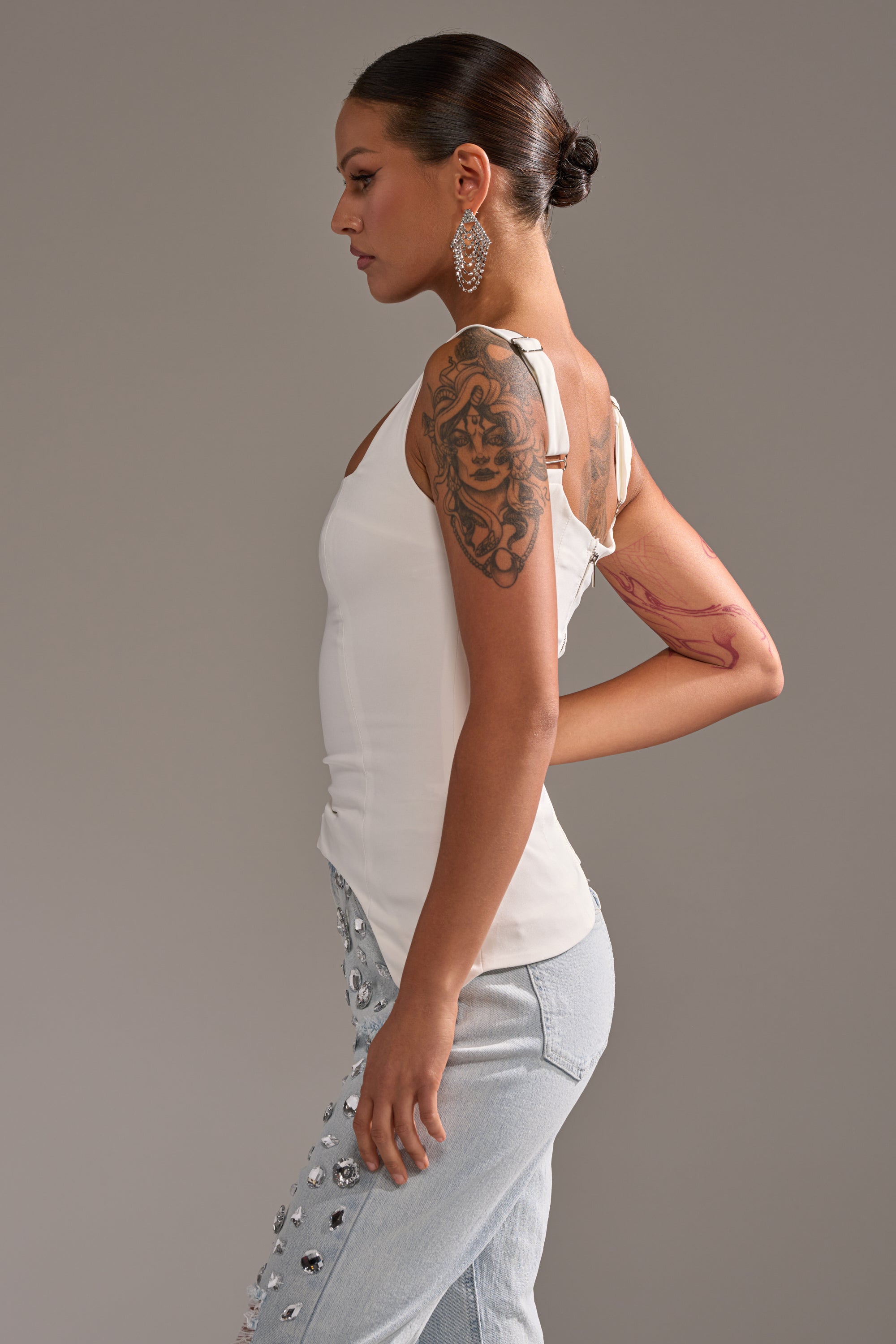 A woman in profile stands against a gray background, wearing the PLAIN JANE SLEEVELESS BLOUSE with a square neckline and light blue embellished jeans. Her hair is in a bun, she wears large earrings, and has a detailed tattoo on her upper right arm. 2