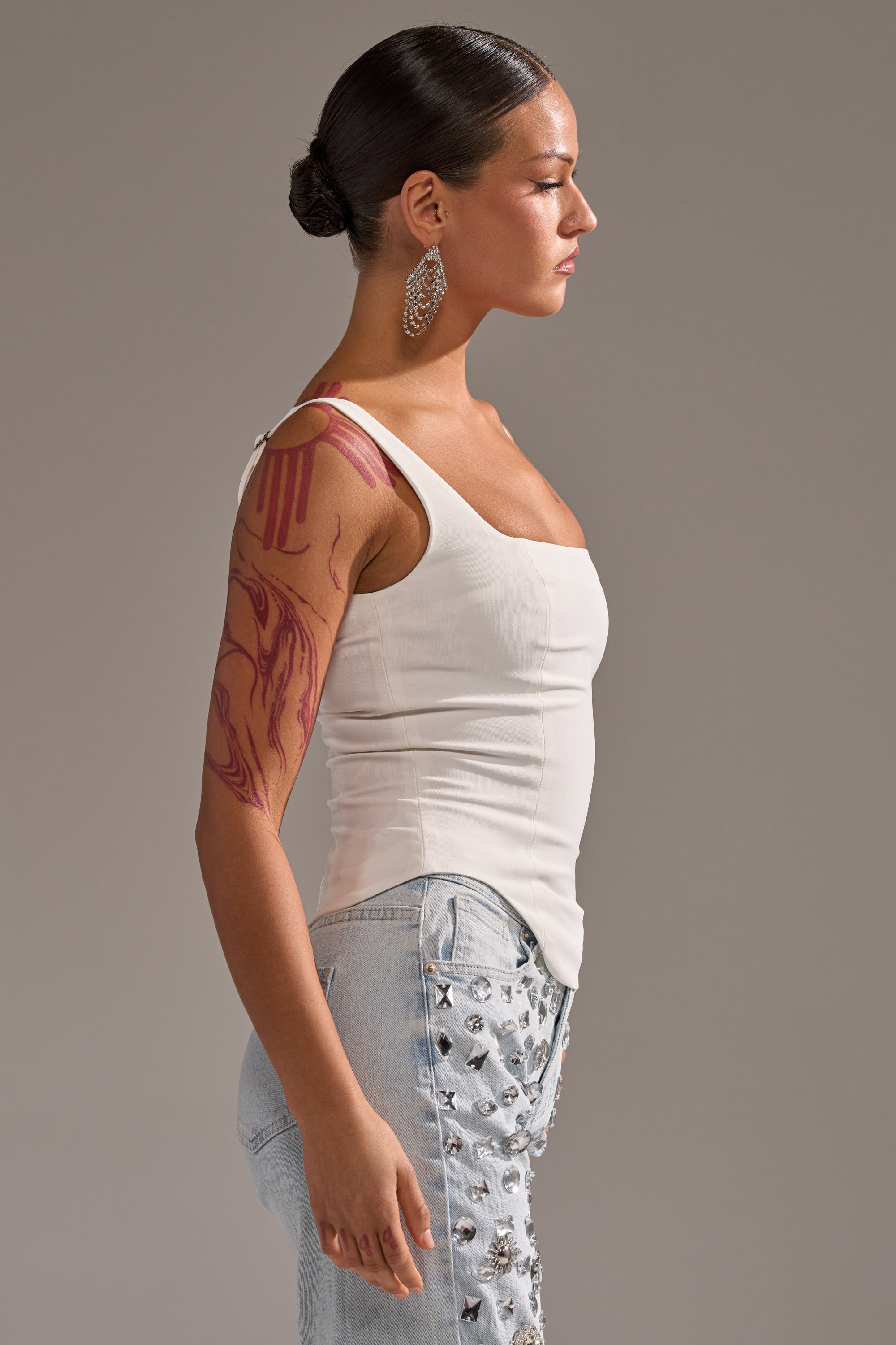 A woman with dark hair in a low bun wears the PLAIN JANE SLEEVELESS BLOUSE, a white square neck top, paired with light blue embellished jeans. She accessorizes with large hoop earrings and has a red tattoo on her upper arm, standing in profile. 3
