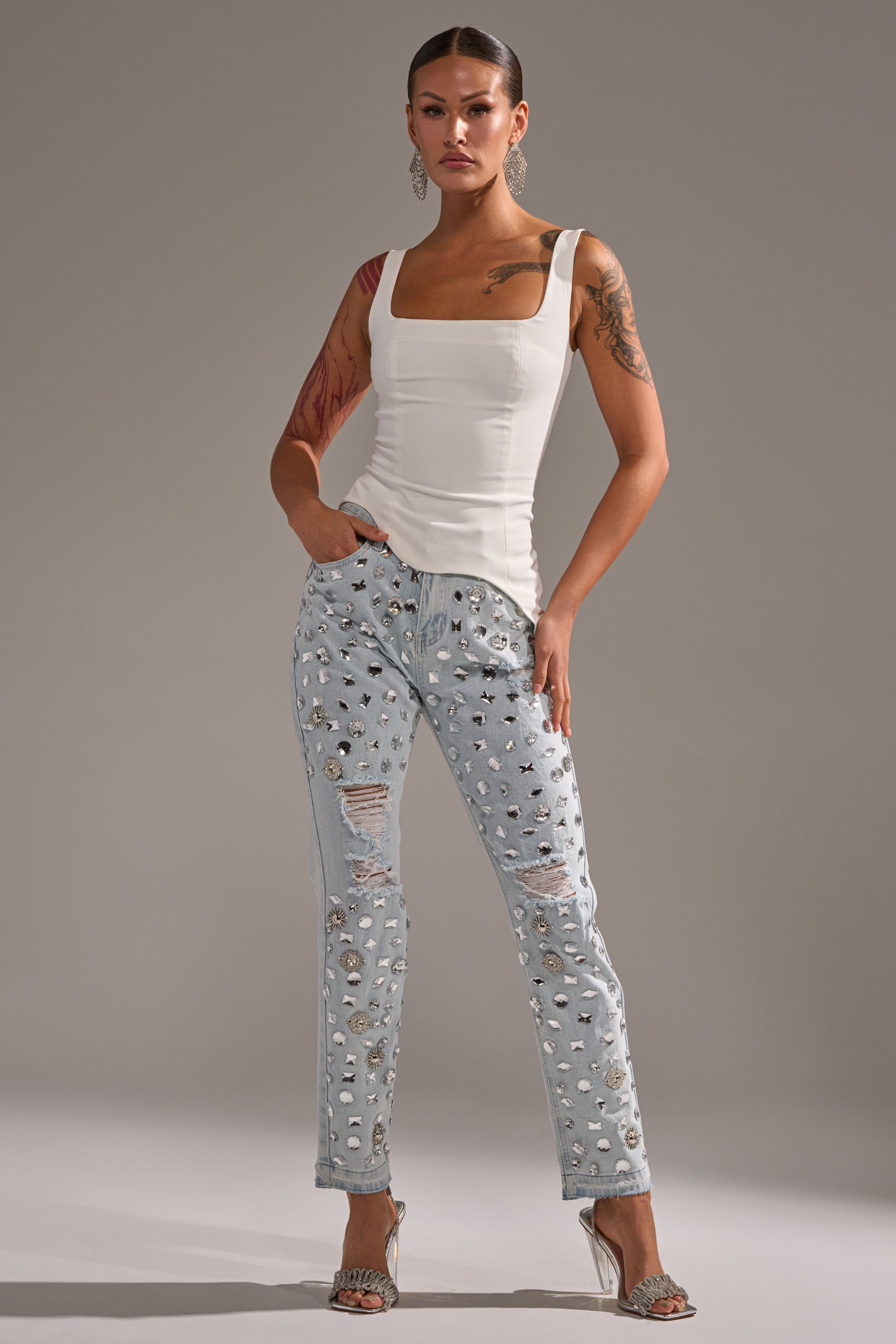 A woman stands against a gray background, wearing the PLAIN JANE SLEEVELESS BLOUSE, light blue ripped jeans with silver embellishments, silver high-heeled sandals, large earrings, and her hands in her pockets. 6