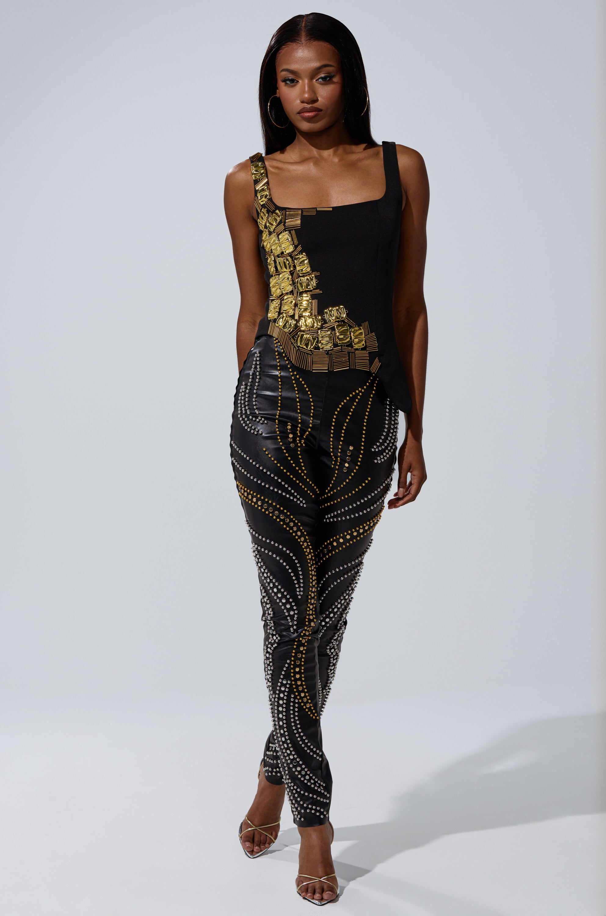 A woman models the HAND BEADED LUX BLOUSE, an asymmetrical black top with gold hardware and beadwork, paired with black pants adorned with swirling gold and silver studs and clear heels against a plain gray background.