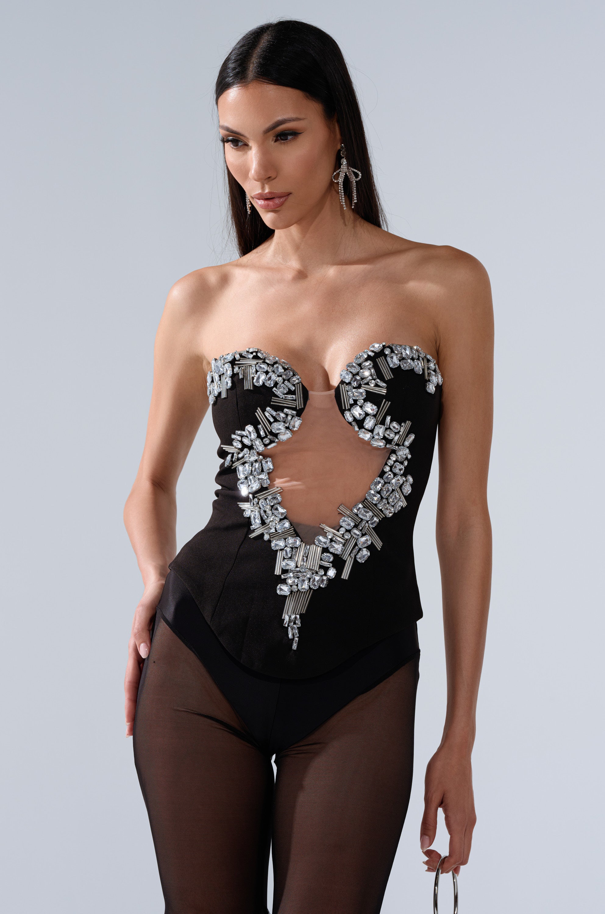 A woman stands against a plain gray background wearing the SHINE ON ME RHINESTONE CORSET, a strapless black top with silver embellishments and a deep V cutout, paired with sheer black pants. 0