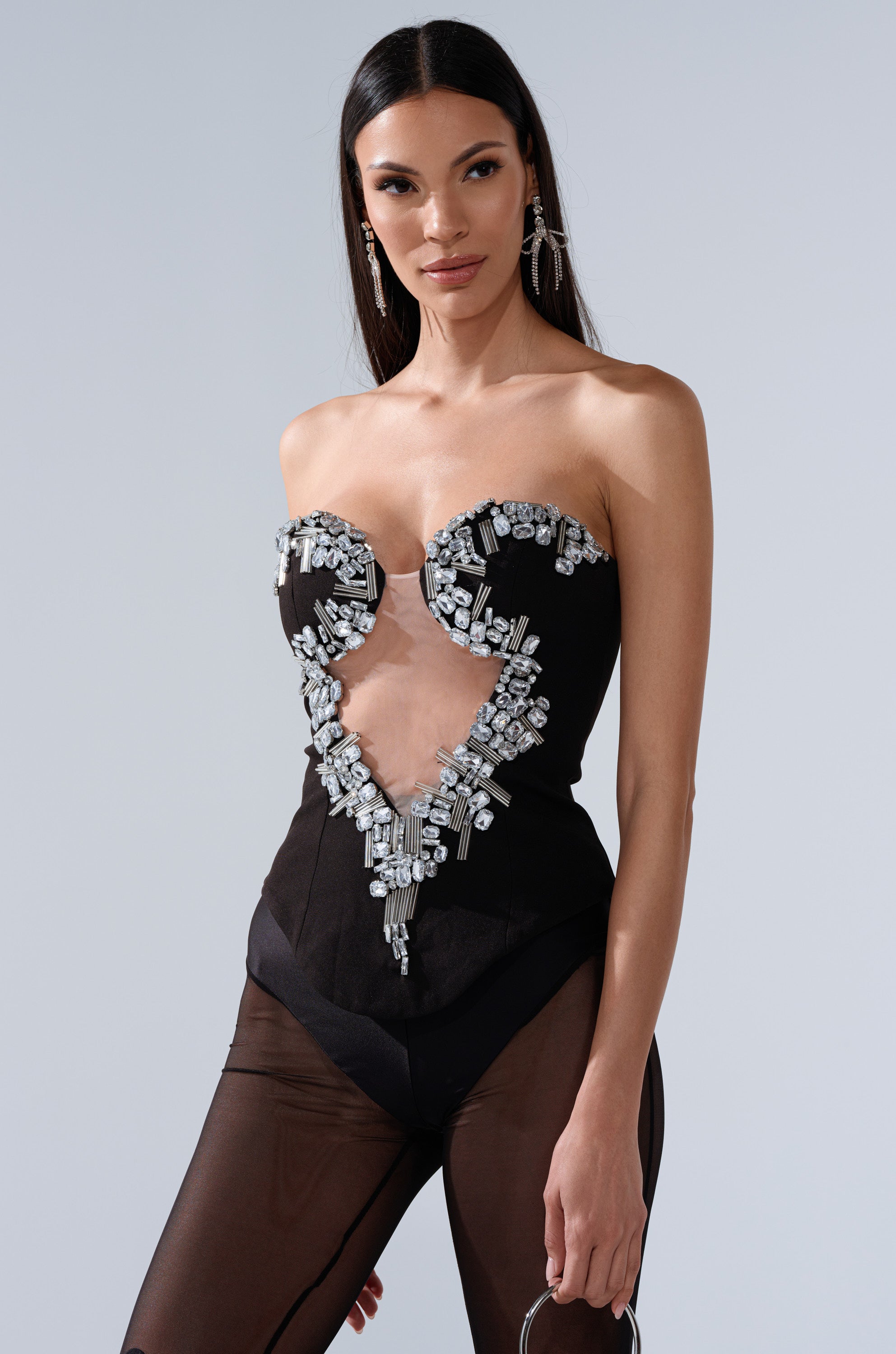 A woman with long, straight dark hair wears the SHINE ON ME RHINESTONE CORSET, a strapless black piece with a sweetheart neckline, sheer V-shaped mesh cut out, and sparkling silver embellishments. She stands against a plain light gray background, smiling. 1