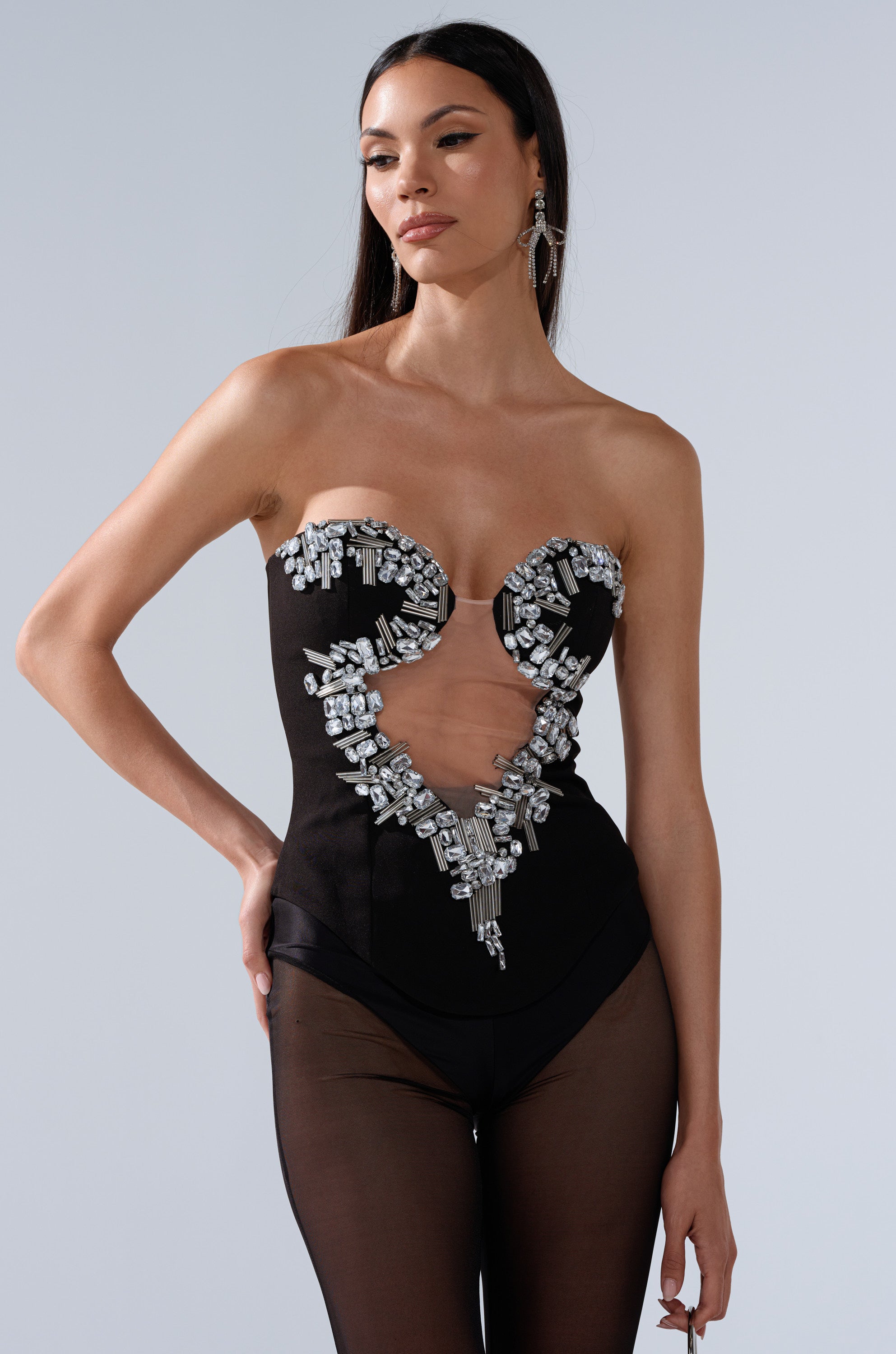 A woman models the SHINE ON ME RHINESTONE CORSET, a strapless black bodysuit with a sweetheart neckline, sparkling rhinestone embellishments, and elegant mesh cutouts. She stands against a plain background with her long dark hair down. 2