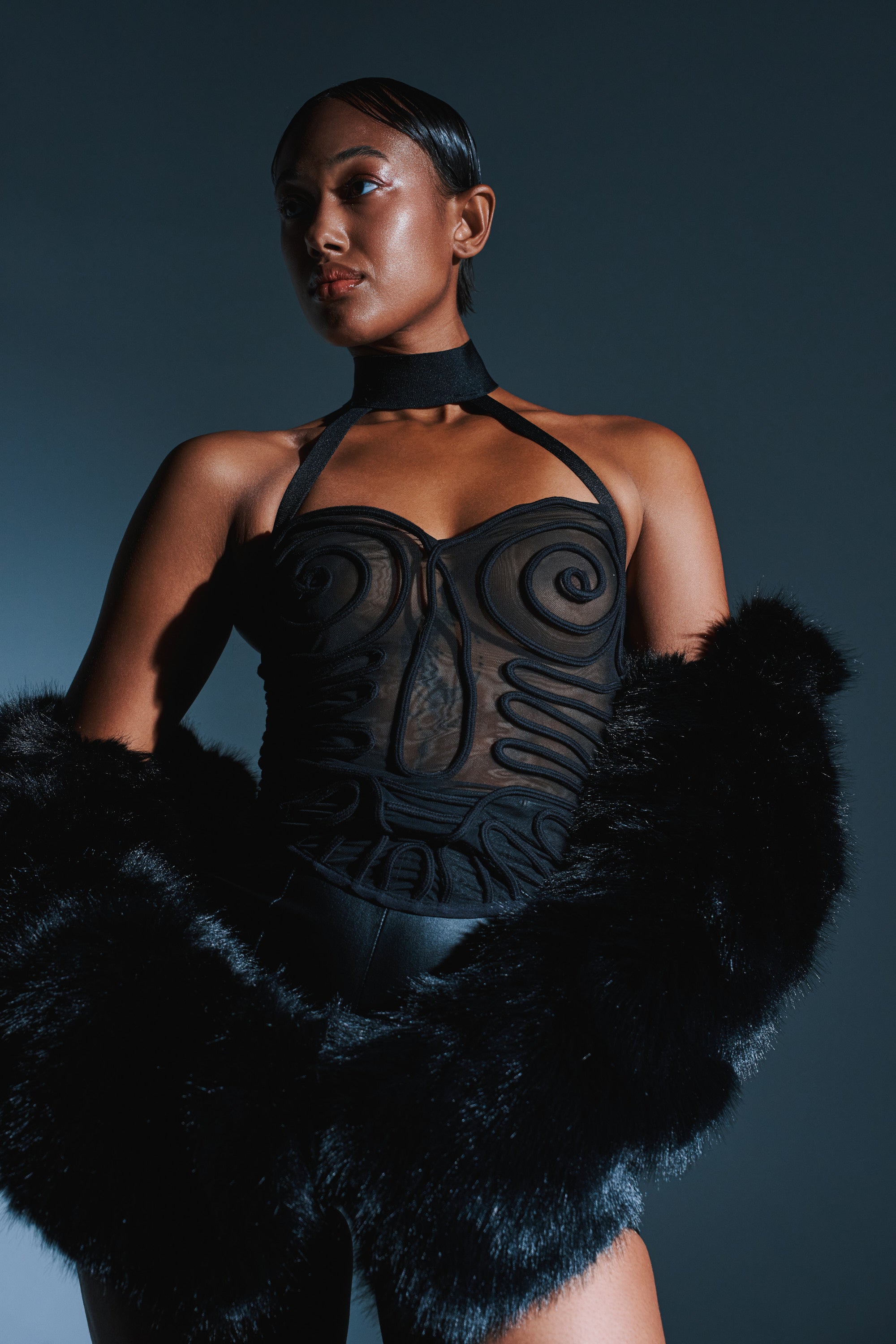A woman poses confidently in the I KNOW THAT’S RIGHT MESH CORSET, featuring a fitted, sheer black mesh design with swirl paneling and a halter neckline. She stands against a dark blue background with slicked-back hair and a black stole.
