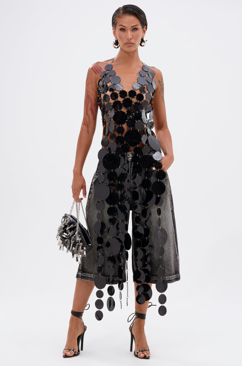 A person stands confidently in the GLAMOROUS CHAIN DETAIL HALTER TOP, paired with cropped faded black denim pants, a small handbag, and black high-heeled sandals. Their hair is slicked back in a wet-look style.