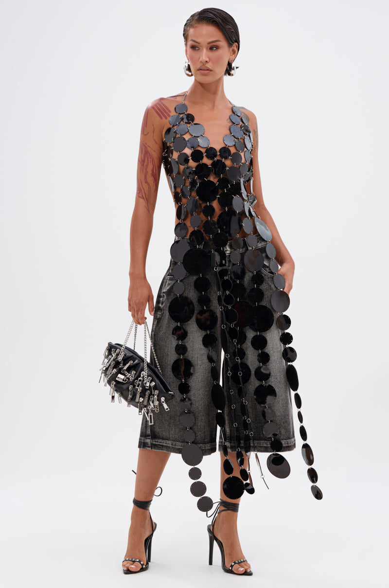 A woman exudes confidence in the GLAMOROUS CHAIN DETAIL HALTER TOP, paired with faded wide-leg jeans. She accessorizes with a studded handbag, high-heeled sandals, has slicked-back hair, and displays visible arm tattoos.