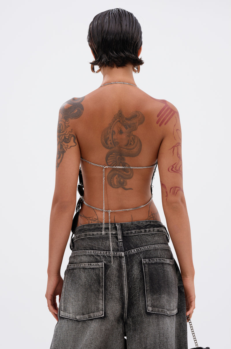 A person with short, dark slicked-back hair models the GLAMOROUS CHAIN DETAIL HALTER TOP and loose, faded black jeans, revealing tattoos on their back and arms while standing against a plain white background.