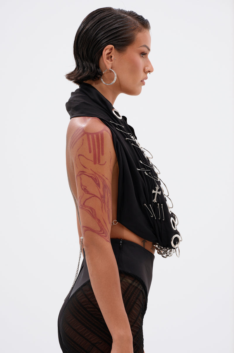 A person with short, slicked-back hair stands in profile, wearing the OVER EMBELLISHED HALTER TOP featuring large metal rings and chains, paired with sheer black pants, bold hoop earrings, and a striking red abstract tattoo on their upper arm. 2
