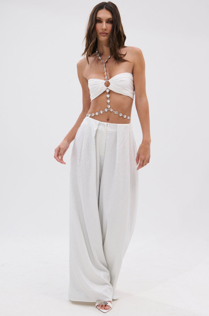 A woman poses in the NOT FEELING IT BRALETTE, white wide-leg pants, and open-toe heels, styled with a beaded heart chain harness against a plain white background.