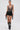 A woman stands against a plain background wearing the CHAINED DOWN HALTER NECK BLOUSE in gunmetal micro-sequins, black leather fringe shorts, chain-trimmed black cowboy boots, hoop earrings, and her hair tied back.