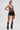 A woman poses in a sleeveless CHAINED DOWN HALTER NECK BLOUSE made from metallic mesh, paired with black fringe leather shorts and tall chain-embellished boots, set against a plain white background.
