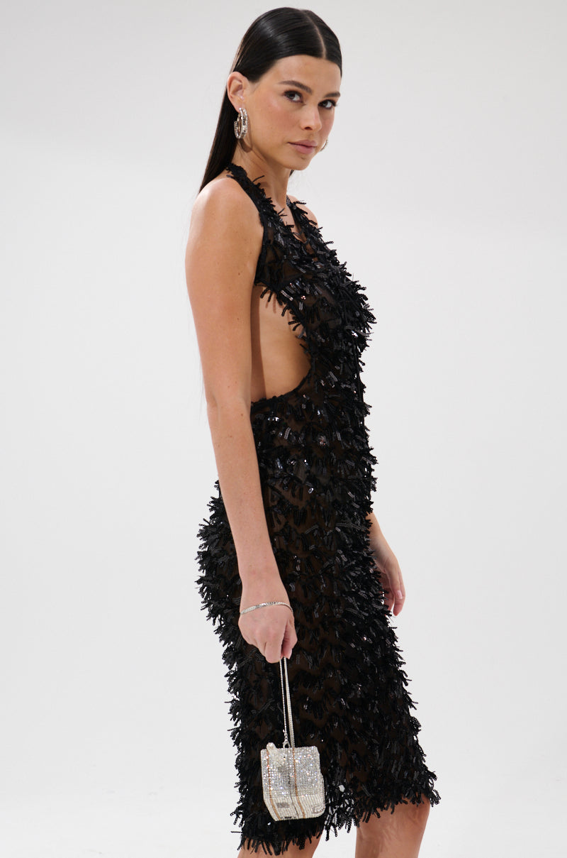 A woman with long dark hair models the LENNOX SEQUIN MESH MIDI DRESS, a curve-hugging black textured dress with a side cutout. She holds a silver purse and poses against a plain white background.