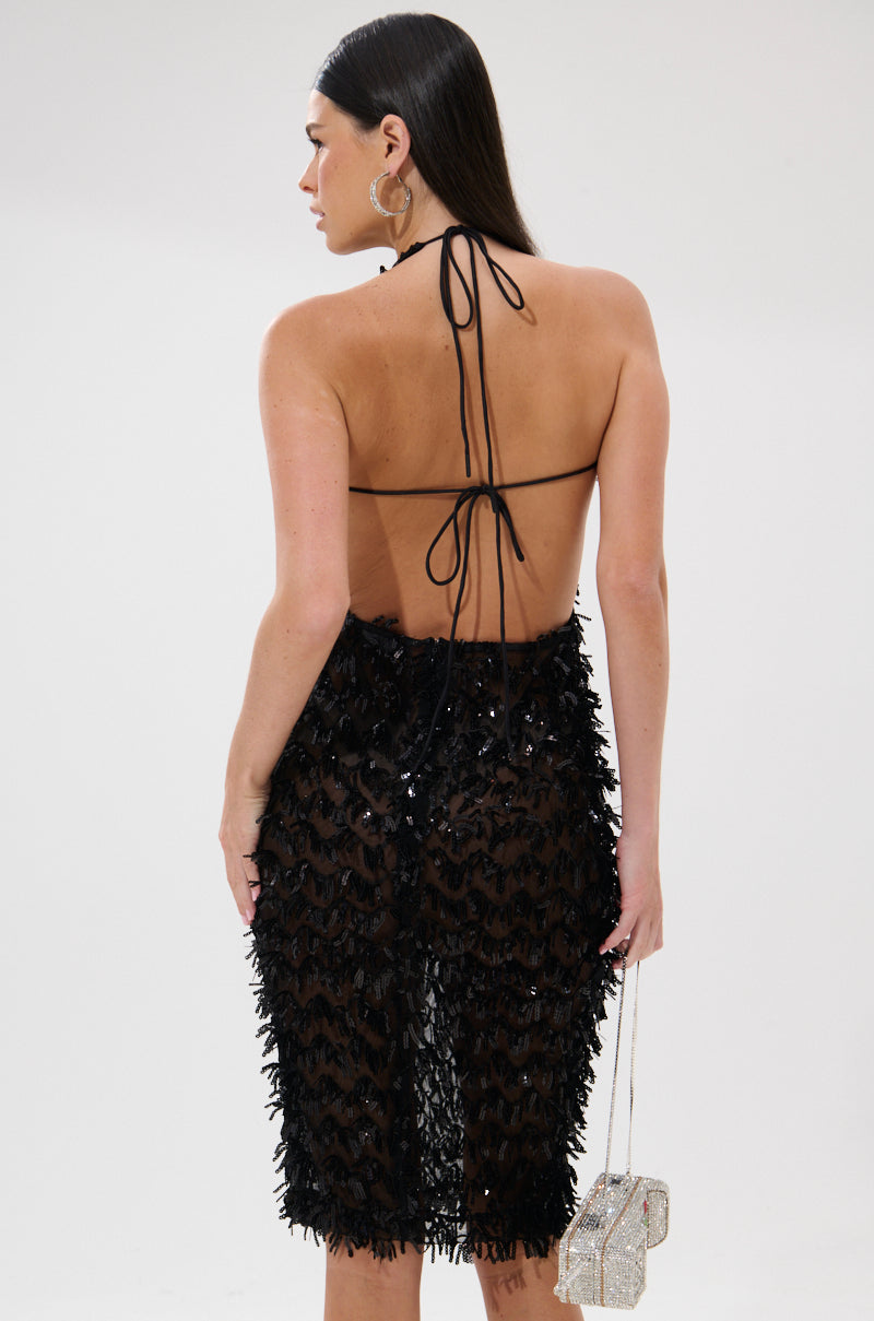 Woman faces away from the camera, wearing the LENNOX SEQUIN MESH MIDI DRESS—a curve-hugging black halter-neck with open back and fringe. She holds a sparkly handbag and has long, straight dark hair.