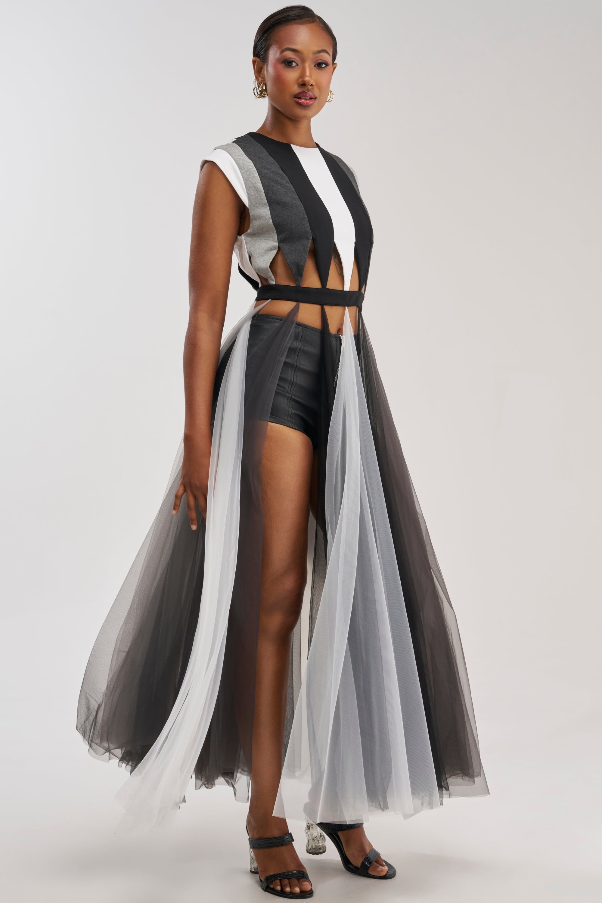 A woman models the FEELING THE MOTIONS TOP—a sleeveless, colorblocked tulle piece with black-and-white geometric patterns and sheer, flowing panels—paired with black shorts and heels as she strikes a confident pose against a light gray background, epitomizing bold editorial style.